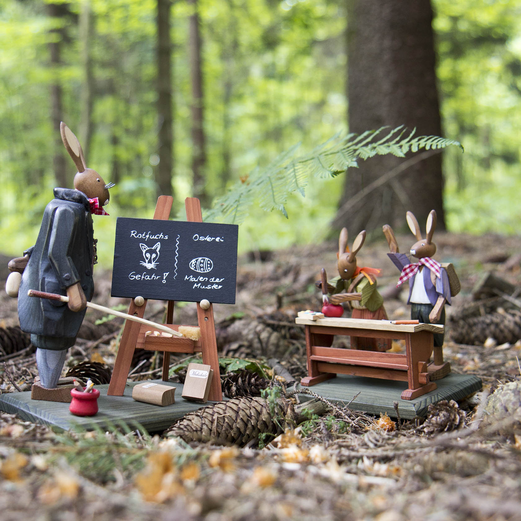 Wooden figurines of rabbits in a forest setting; one rabbit acts as a teacher pointing at a blackboard with drawings, and two other rabbits sit at a desk.