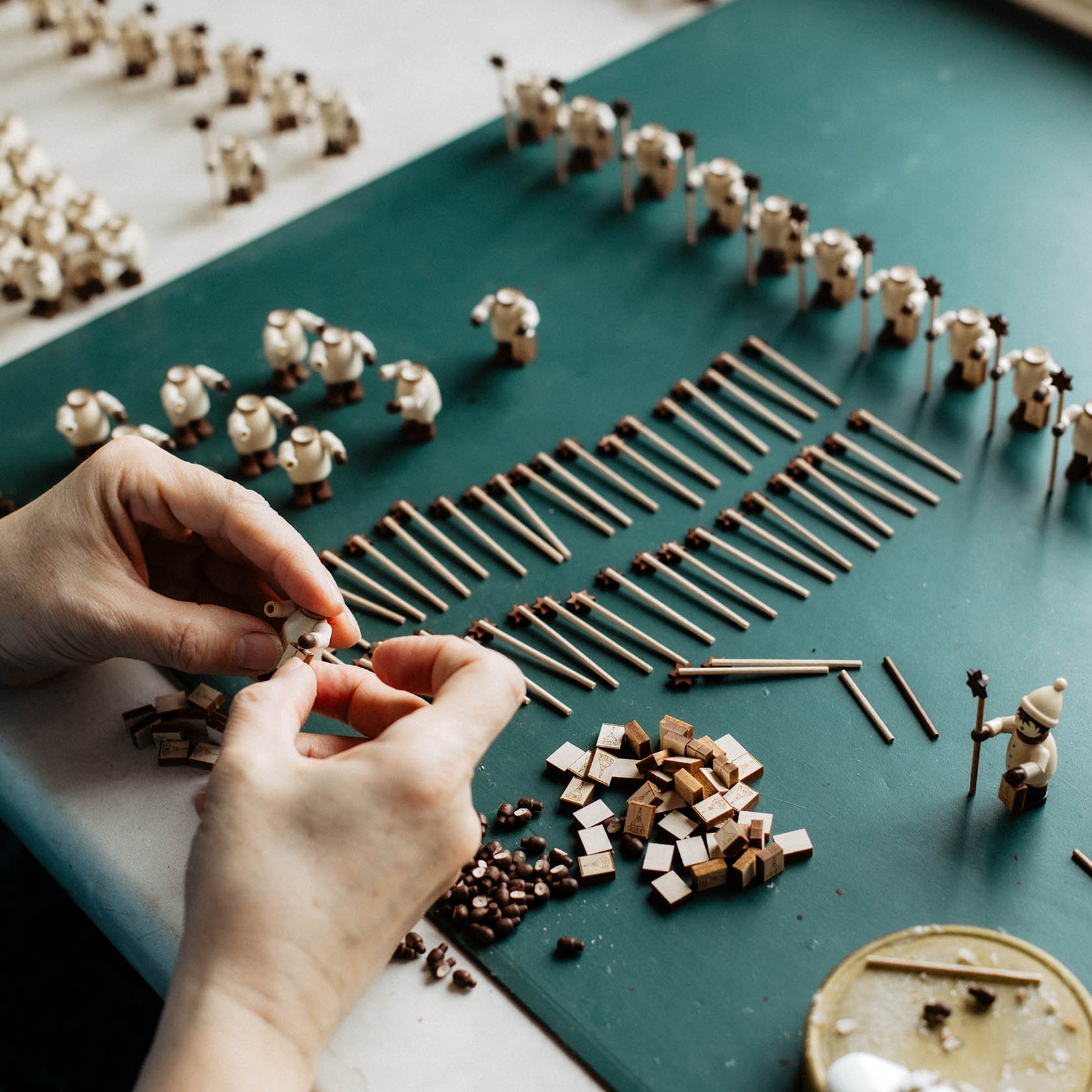 Hands assembling small natural wood Kurrende figures with several parts and completed figures on a green table.