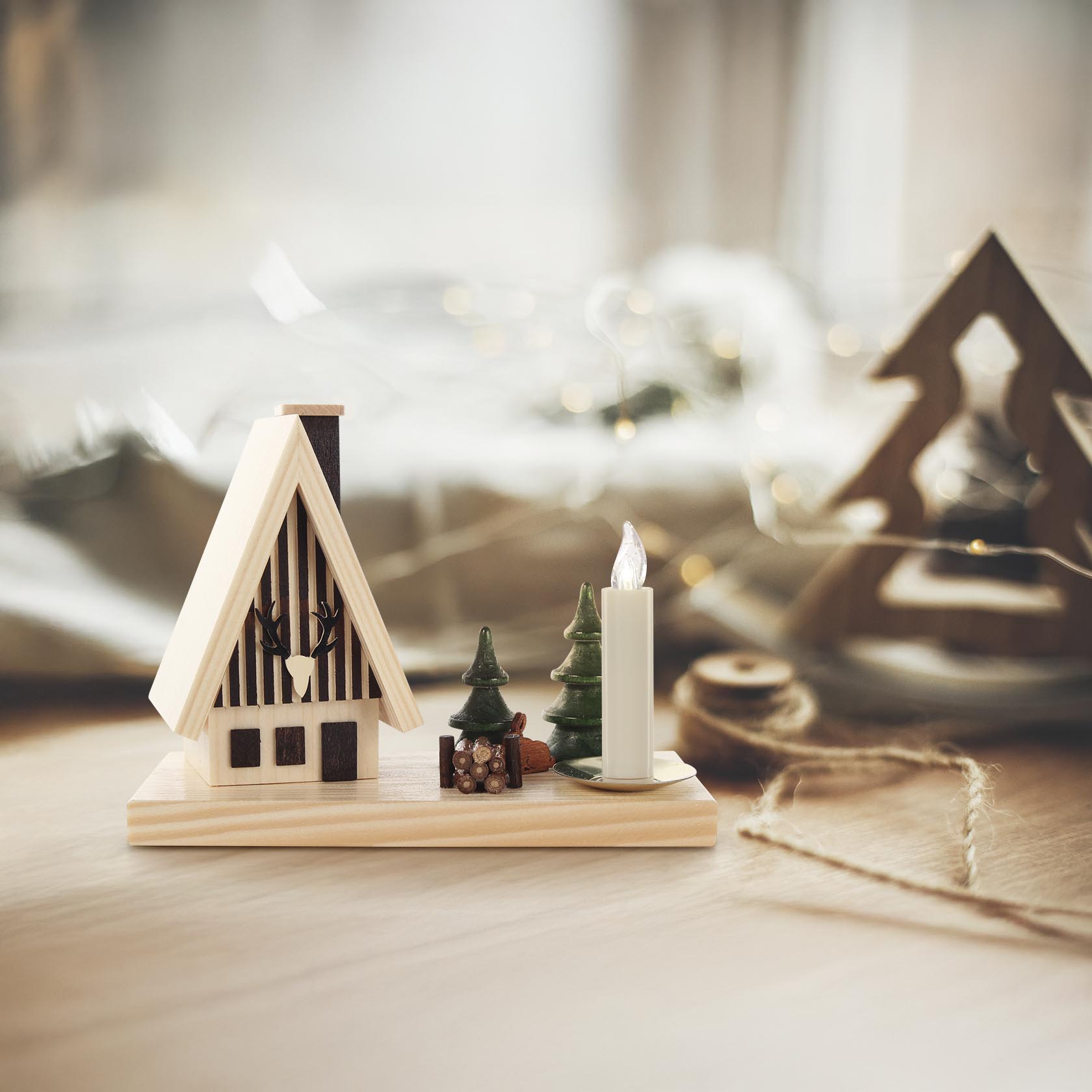 Räucherhaus in Form eines Waldhauses mit Spitzdach, flankiert von einem grünen Baum und einem sitzenden Hasen. Daneben ein Kerzenhalter mit einer weißen Kerze.
