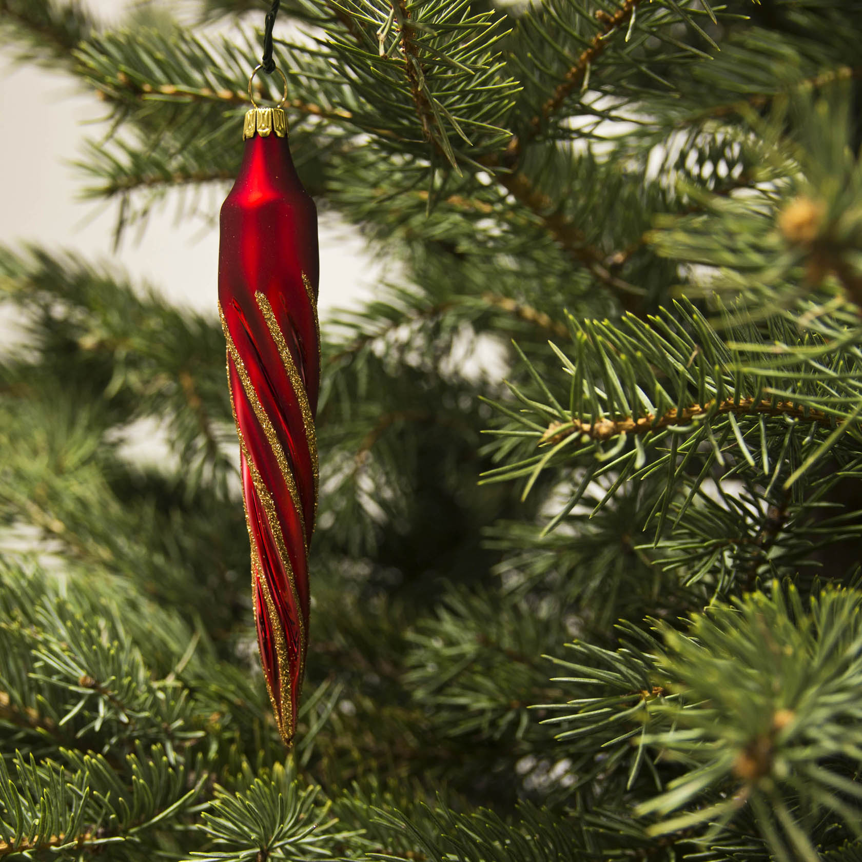 A red matte icicle-shaped Christmas ornament with gold glitter accents hanging on a green Christmas tree branch.