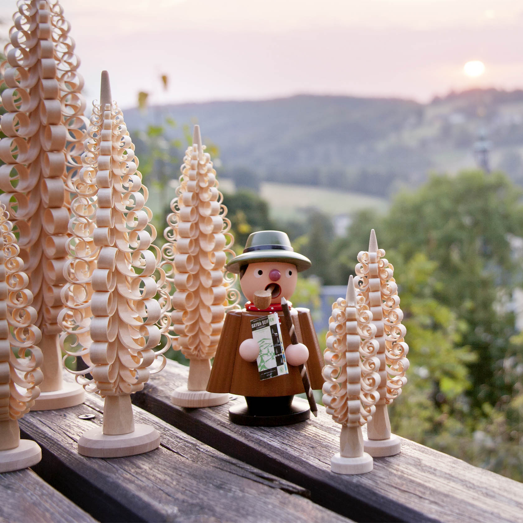 A traditional wooden smoker figurine shaped like a hiker, surrounded by handcrafted wooden trees, set against an outdoor backdrop.
