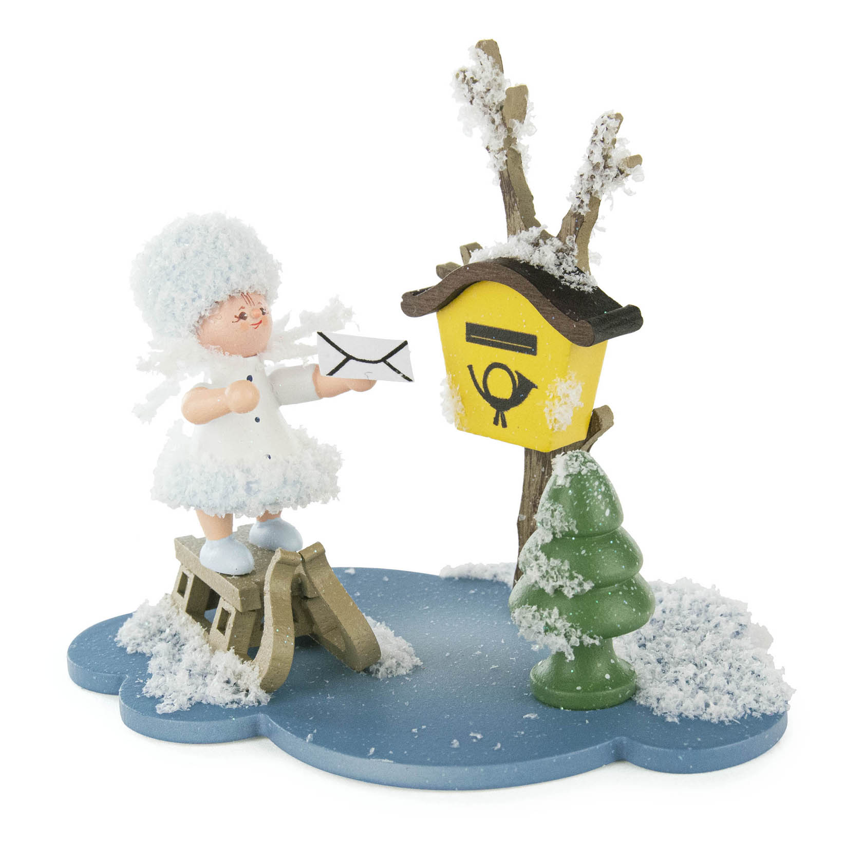 Holzfigur eines Schneeflöckchens in weißem Kleid und Mütze, das einen Brief in einen gelben Briefkasten mit Schnee einwirft, daneben ein schneebedeckter Baum.