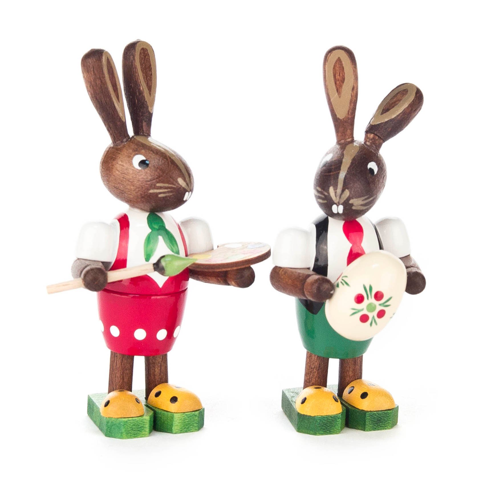 Two wooden bunny figurines, each holding a brush and an egg. One wears a red outfit and the other a green outfit. Both have yellow shoes. im Dregeno Online Shop günstig kaufen