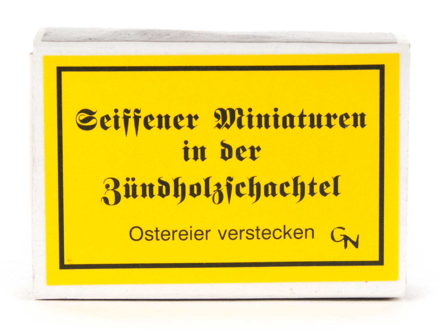 A small box with a yellow label displaying decorative black text that reads 'Seiffener Miniaturen in der Zündholzschachtel' and 'Ostereier verstecken'.