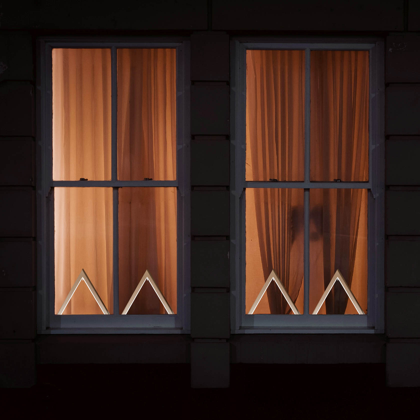 Zwei beleuchtete Fenster mit Vorhängen, in denen jeweils ein beleuchtetes weißes Dreieck mit LED Band zu sehen ist.