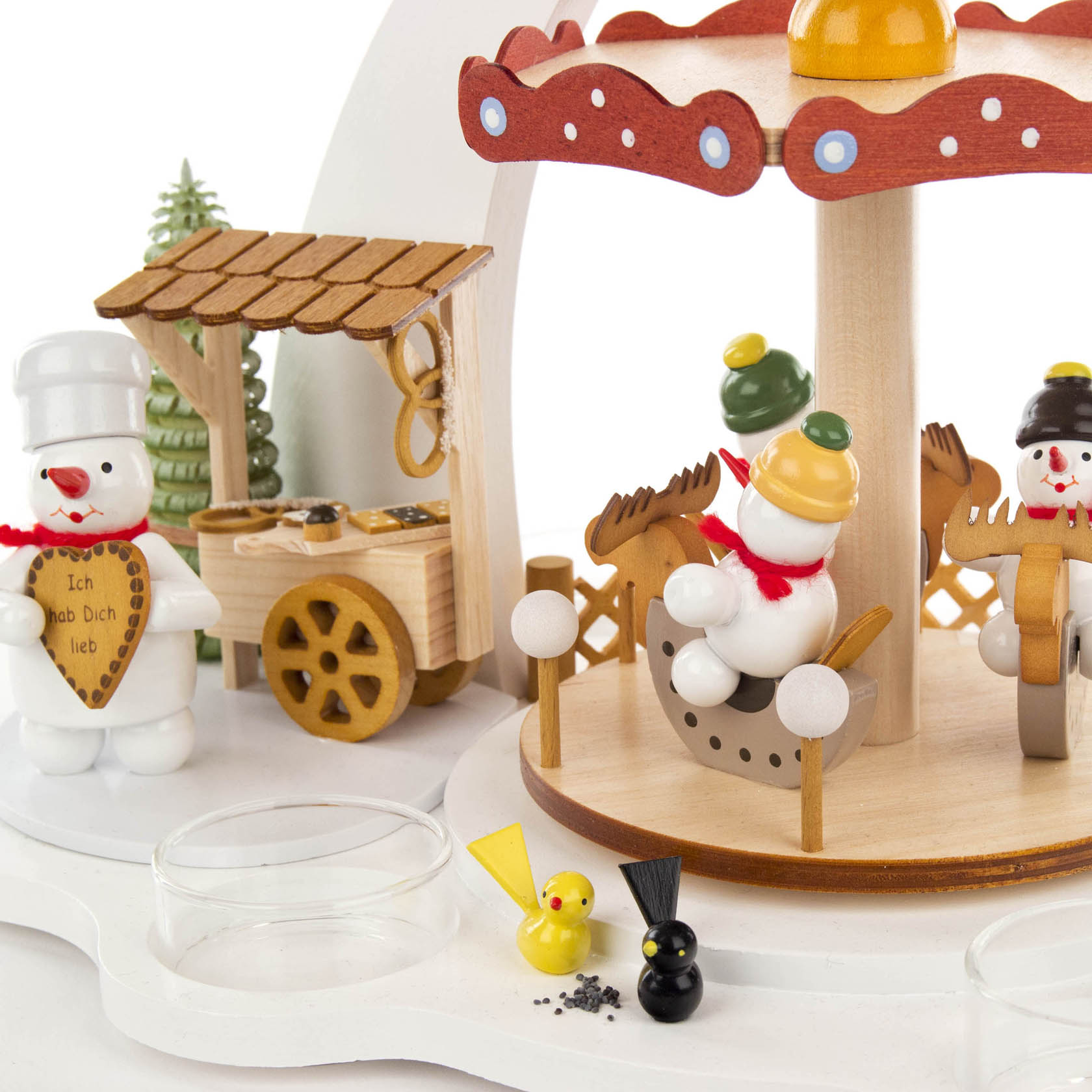 A white pyramid carousel designed for tea lights, featuring snowmen figurines. Some snowmen are riding reindeer on the carousel, another is holding a heart inscribed with 'Ich hab Dich lieb,' standing near a wooden cart.