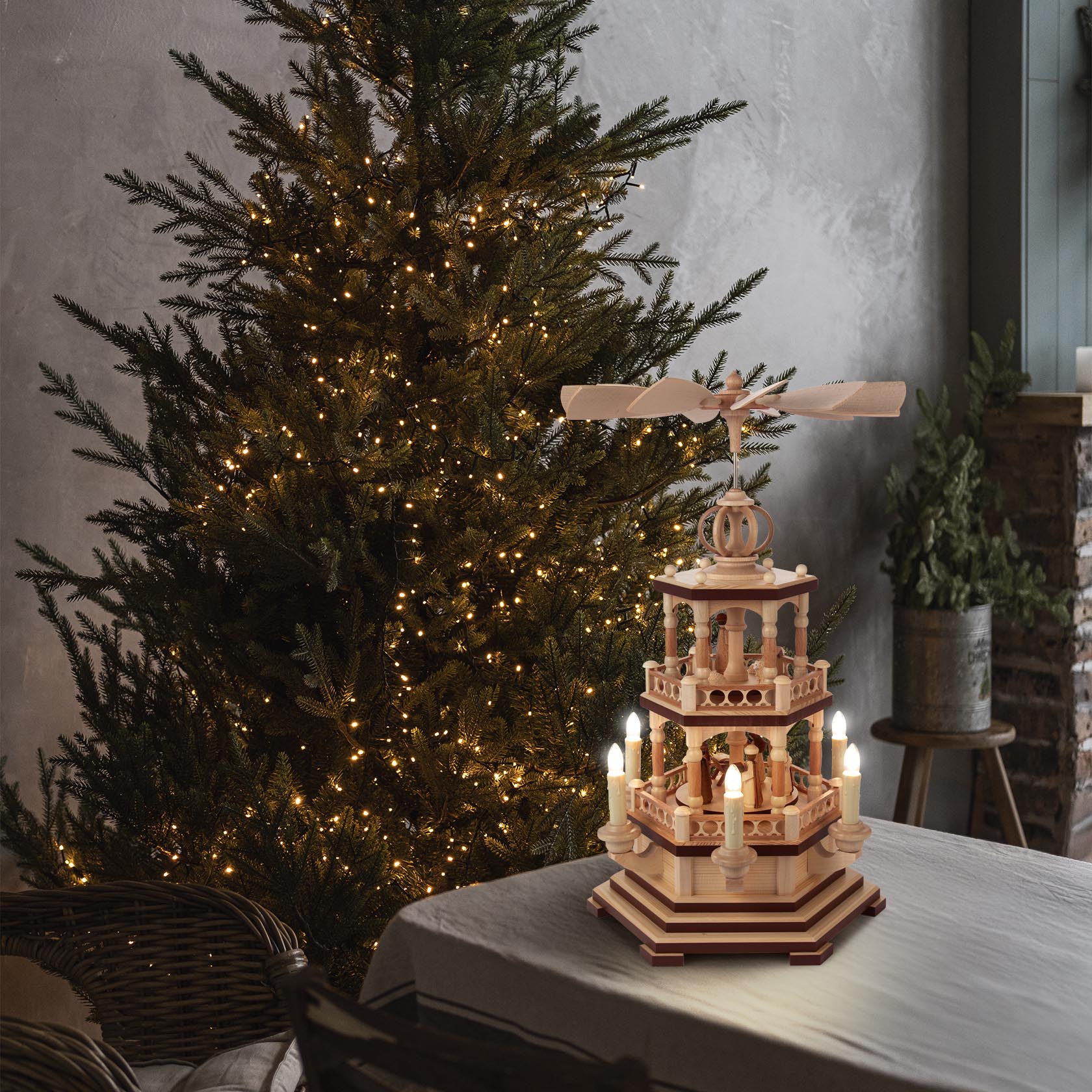 Eine zweistöckige Holzpyramide mit Christi Geburt Szene und elektrischer Beleuchtung auf einem Tisch, im Hintergrund ein beleuchteter Weihnachtsbaum.