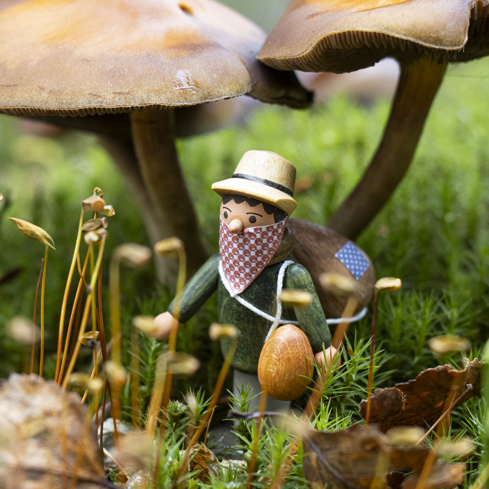 A small wooden figurine of a hiker with a hat and bandana, carrying a wooden stick and a bag, walking among large mushrooms and moss.