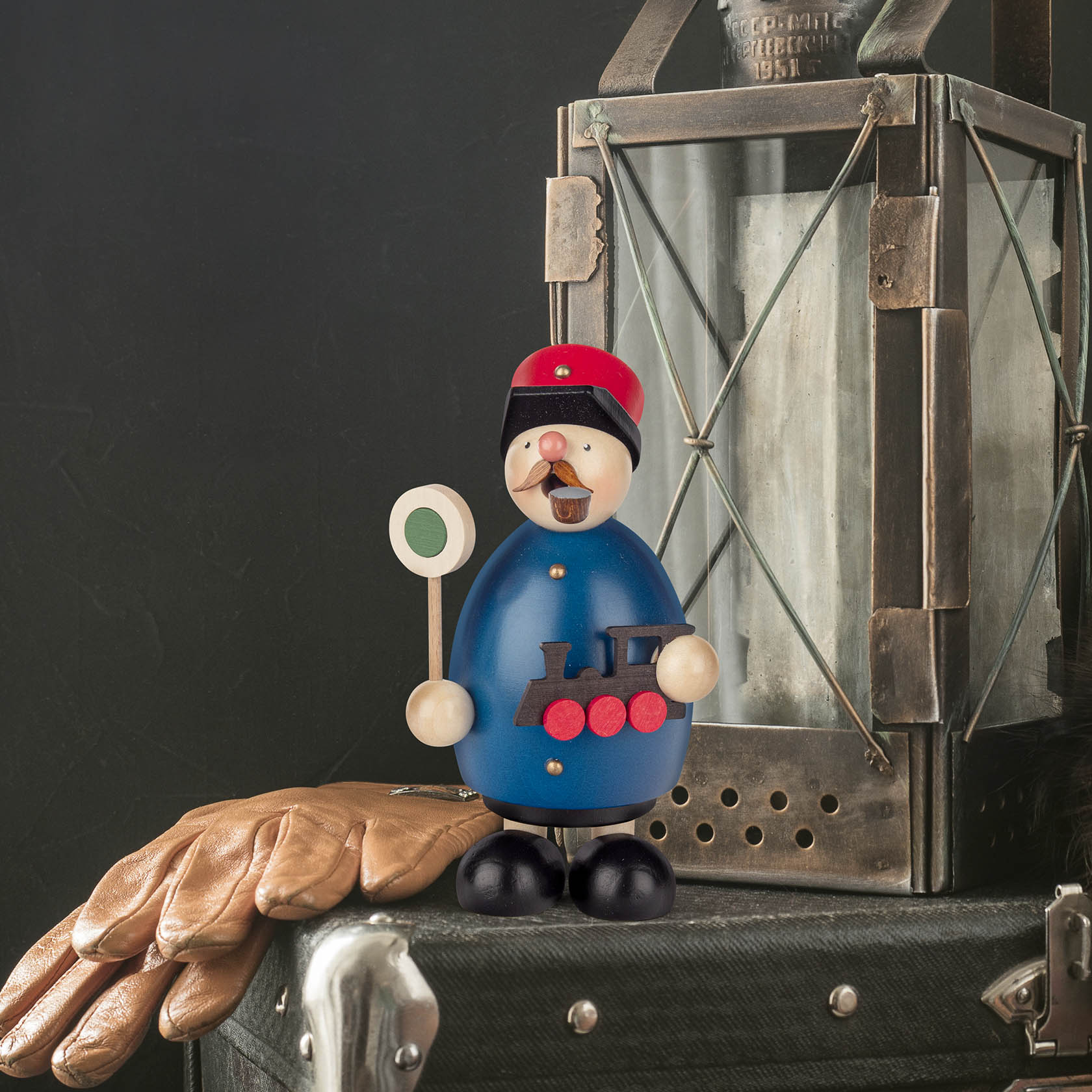 Ein Räuchermann in Eisenbahnerkleidung mit roter Mütze und blauer Jacke, darauf eine schwarze Lokomotive. Er hält ein rundes Schild in der Hand.