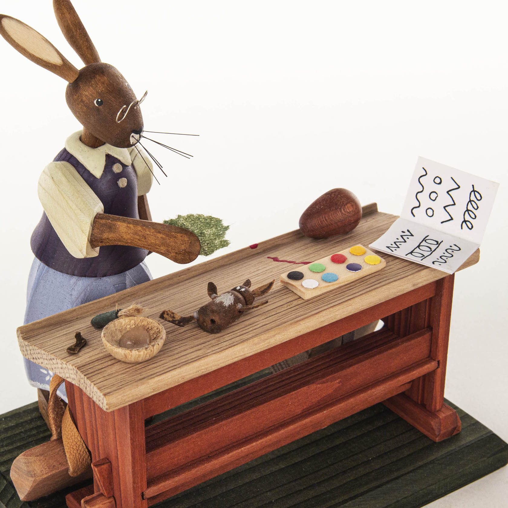 Wooden bunny figure dressed in a blue outfit, standing at a school desk with a small easter egg, paint palette, and painted line patterns on paper.