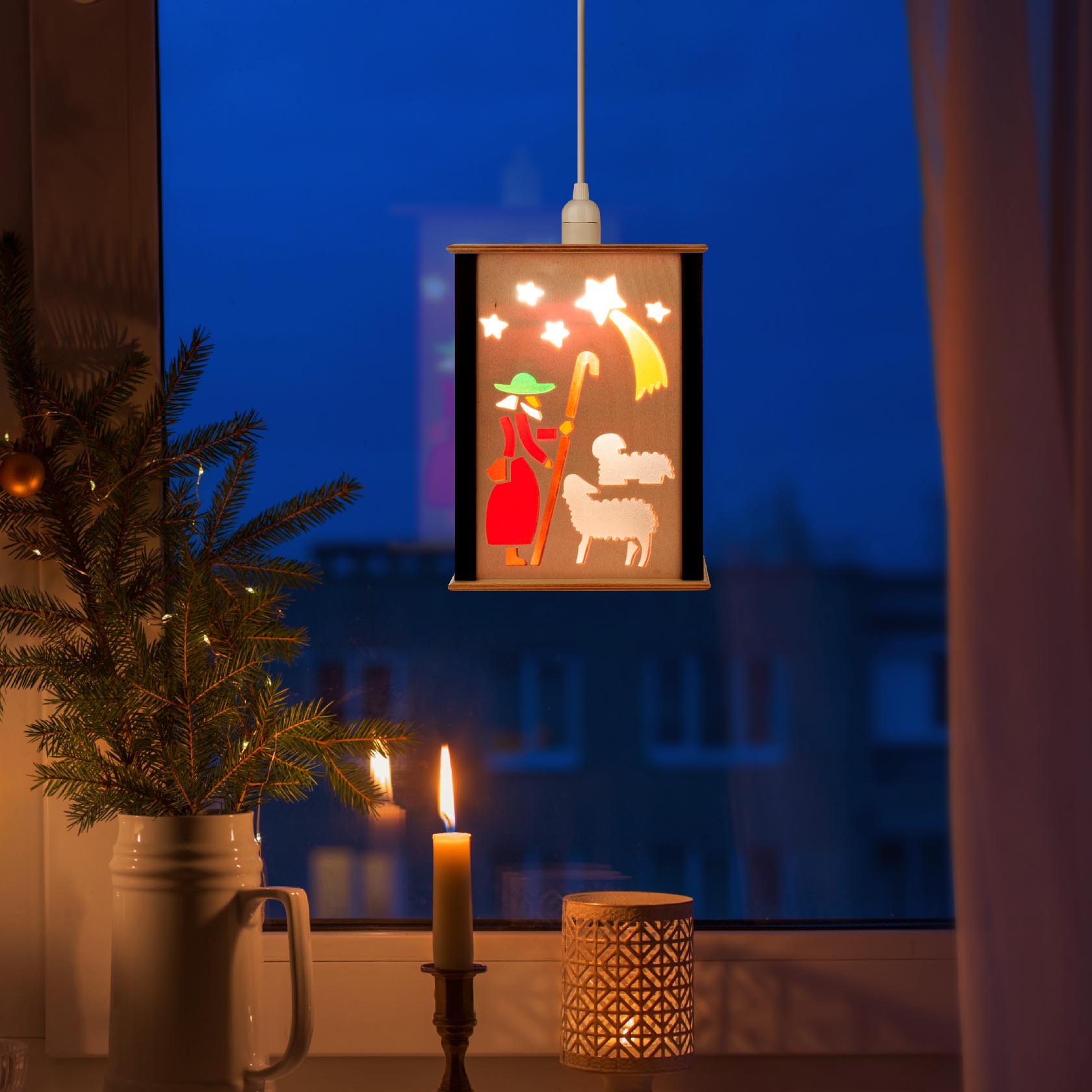 A small window lantern depicting the Nativity scene with a shepherd, a sheep, and a bright star. It's hanging in a warm, cozy room by a candle and a pine branch.