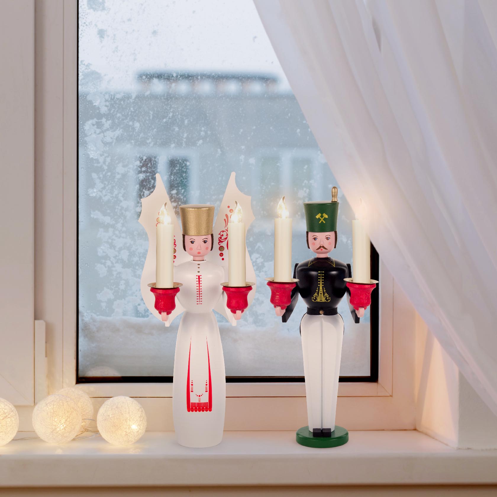 Holzfiguren von Engel und Bergmann für Kerzen auf Fensterbank, hinter einem schneebedeckten Fenster.