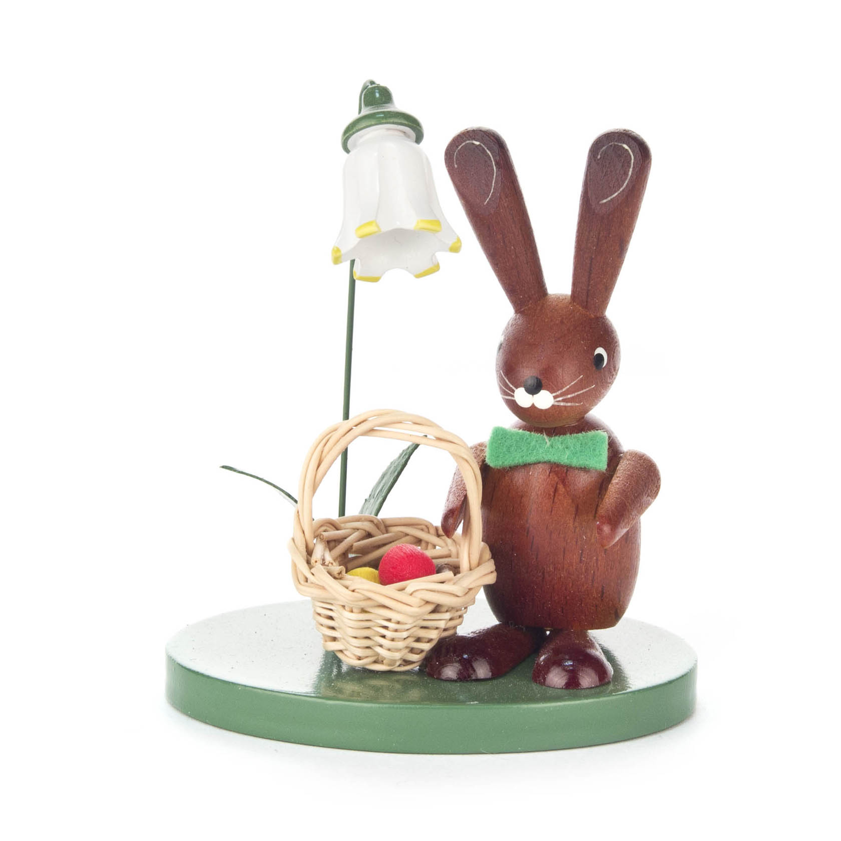 Holzhase mit grüner Fliege, einen Korb mit rotem Ei haltend, neben einer weißen Blume mit gelbem Rand auf einem grünen Sockel.