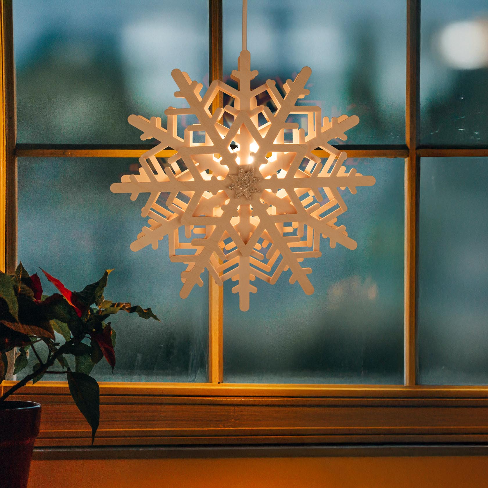 Illuminated snowflake ornament hanging in front of a window, casting a warm light. A plant is in the bottom left corner.