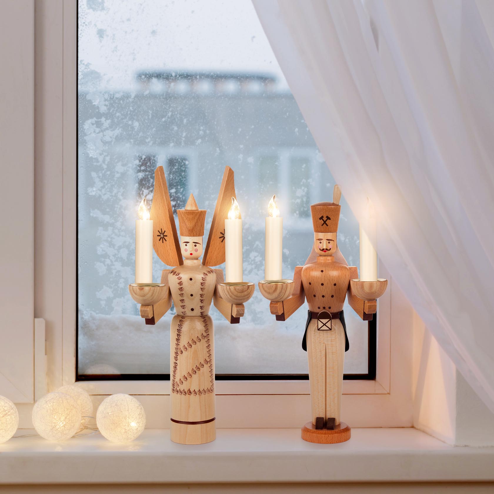 Holzfiguren eines Engels und eines Bergmanns mit Kerzen auf einem Fensterbrett. Der Engel trägt Flügel und eine getupfte Schürze, beide tragen Kerzen.