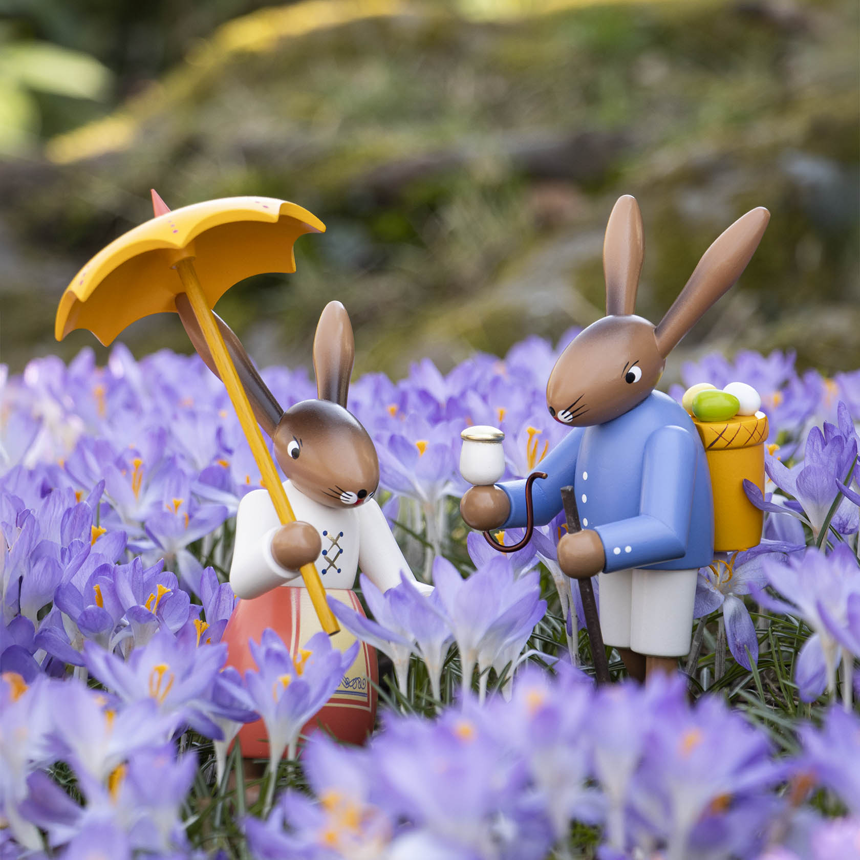 Two Easter bunny figurines in a field of purple flowers. The left bunny holds a yellow umbrella, while the right bunny carries a basket of eggs.
