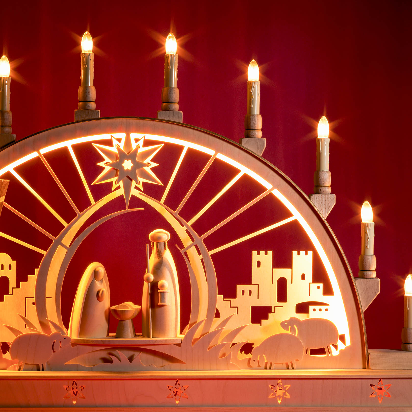 Ein beleuchteter Schwibbogen aus Holz zeigt die Szene der Geburt Christi mit Maria, Josef, dem Jesuskind und Hintergrundbeleuchtung. Kerzen umranden den Bogen.