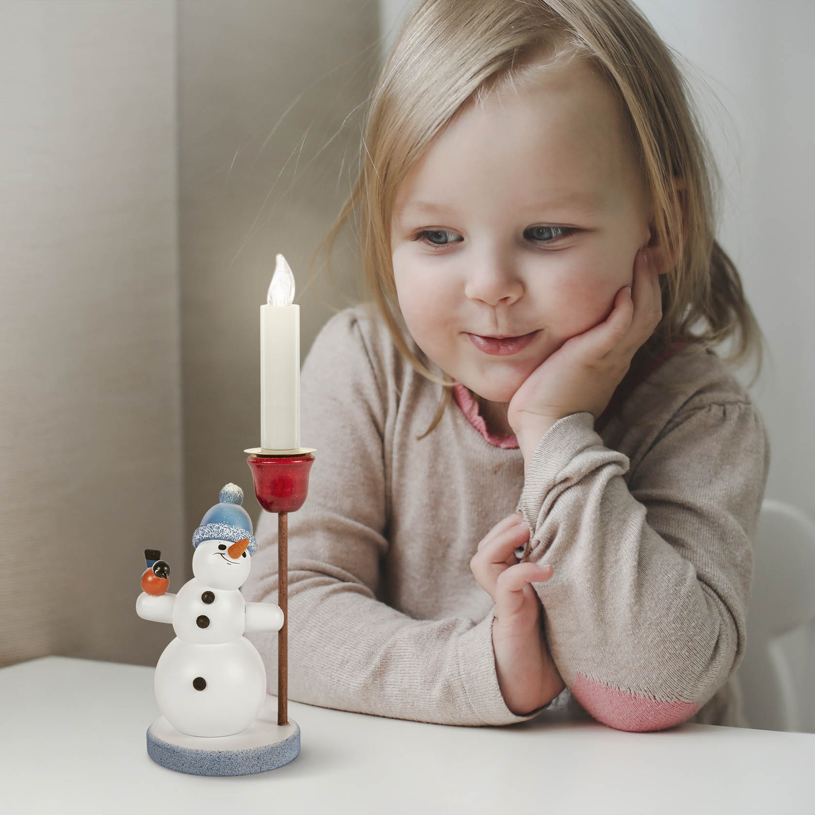 A smiling child looking at a snowman figurine with a blue bobble hat, holding a candle holder for a 14mm diameter candle.