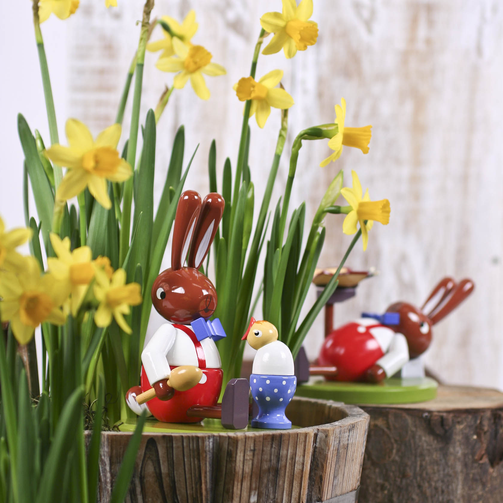 A sitting brown Easter bunny holding a spoon next to a large blue egg cup, surrounded by yellow daffodils.