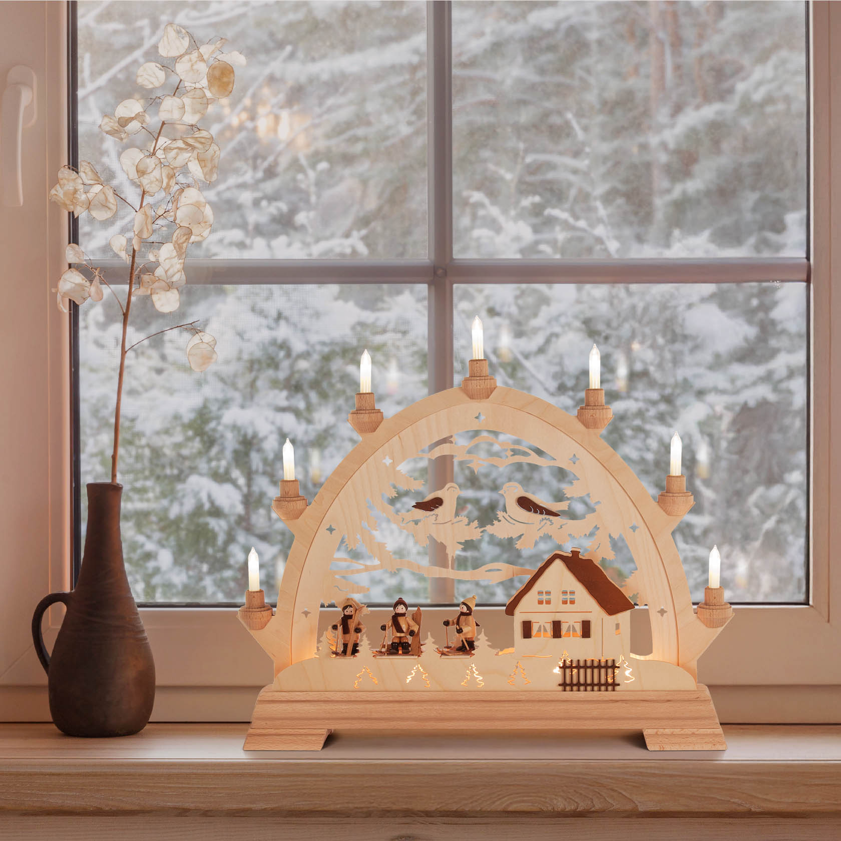 Ein elektrischer Schwibbogen aus Holz, zeigt Skifahrer, ein Haus und Vögel, beleuchtet mit kleinen Kerzen, steht auf einer Fensterbank.