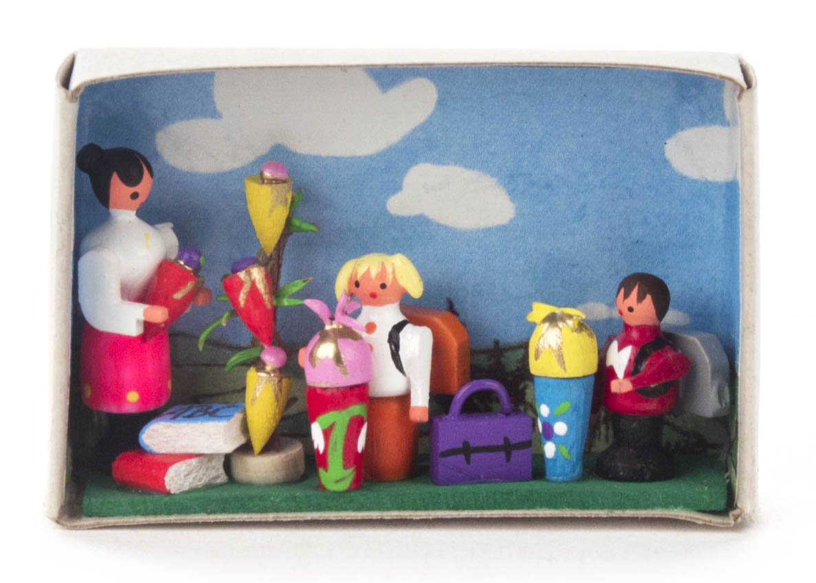A small matchbox diorama depicting a woman and two children at a school start scene. The children hold colorful school cones, symbolizing German school tradition.