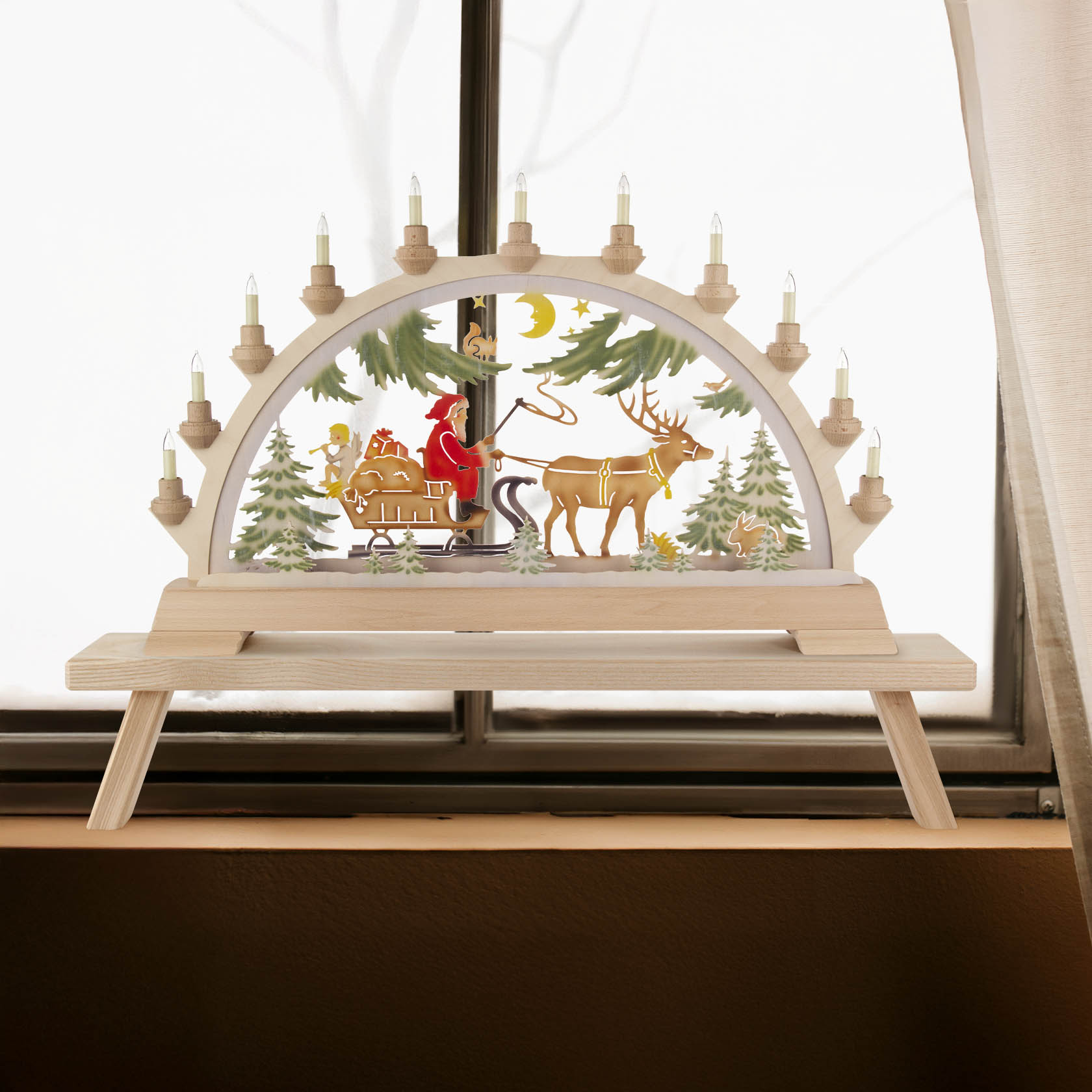 Farbig beleuchteter Schwibbogen mit Nikolaus, der in einem Schlitten mit Geschenken sitzt, gezogen von einem Rentier vor einem verschneiten Waldhintergrund.