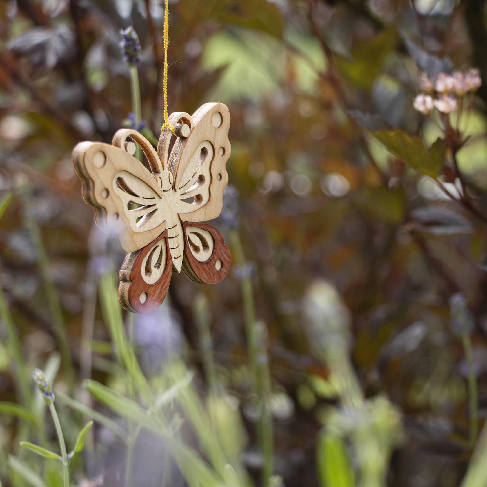 A detailed wooden butterfly ornament hanging amidst lush green foliage and small flowers, showcasing intricate designs and different wood tones.