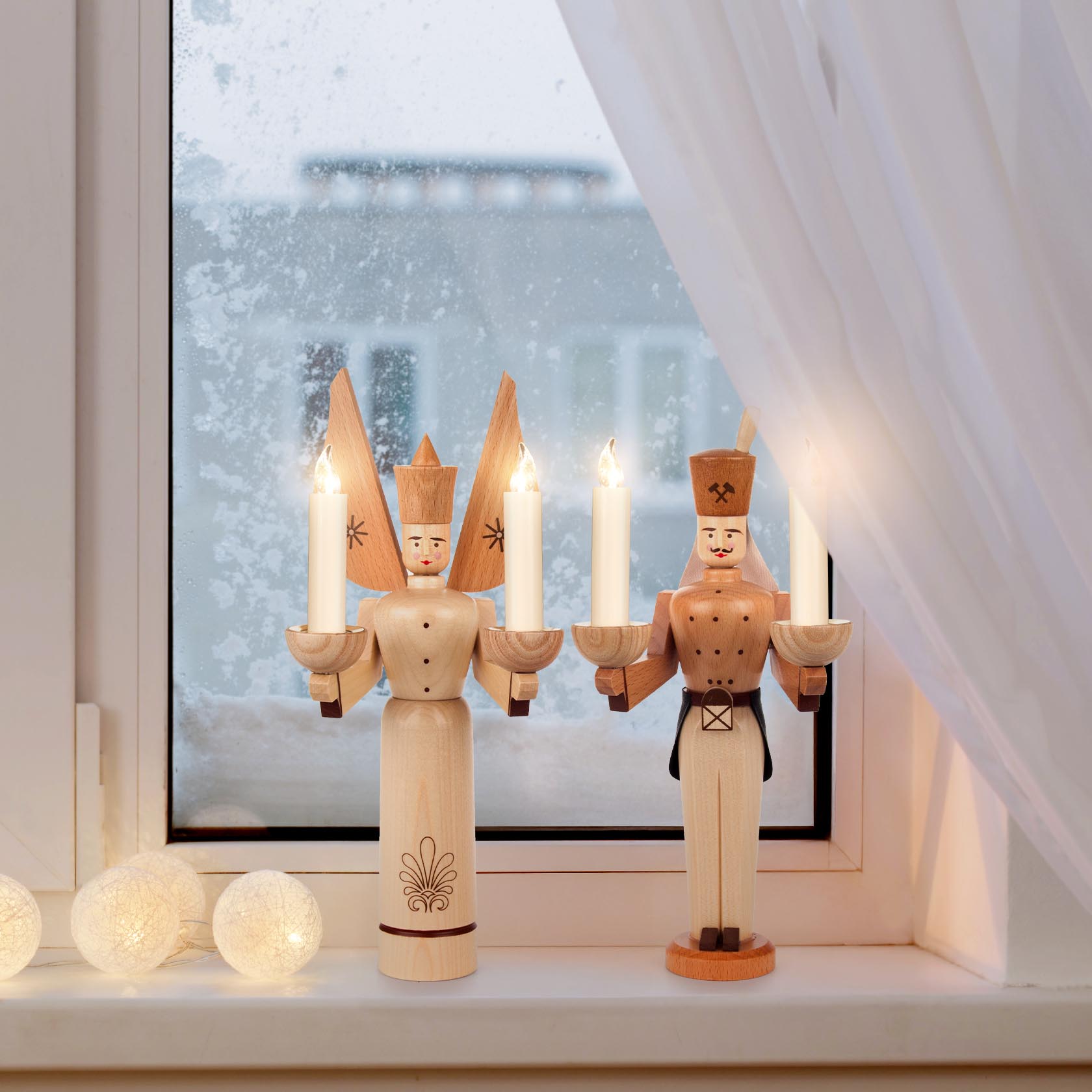 Ein Engel und ein Bergmann aus Holz stehen nebeneinander auf einer Fensterbank. Beide Figuren halten Kerzenhalter mit brennenden Kerzen in den Händen.