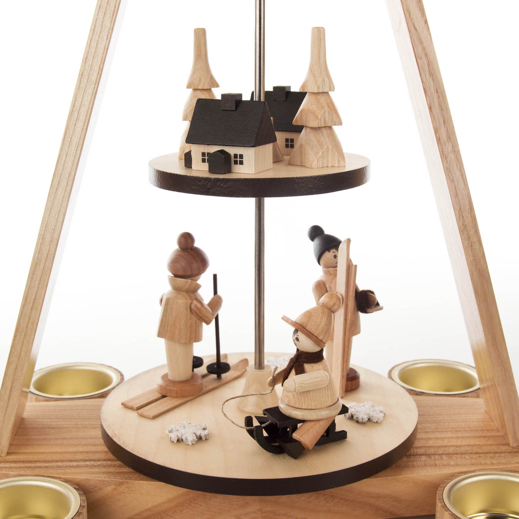 A two-tiered wooden pyramid candle holder with a winter scene. The lower tier features carved figurines of people skiing, and the top tier shows a house and trees.