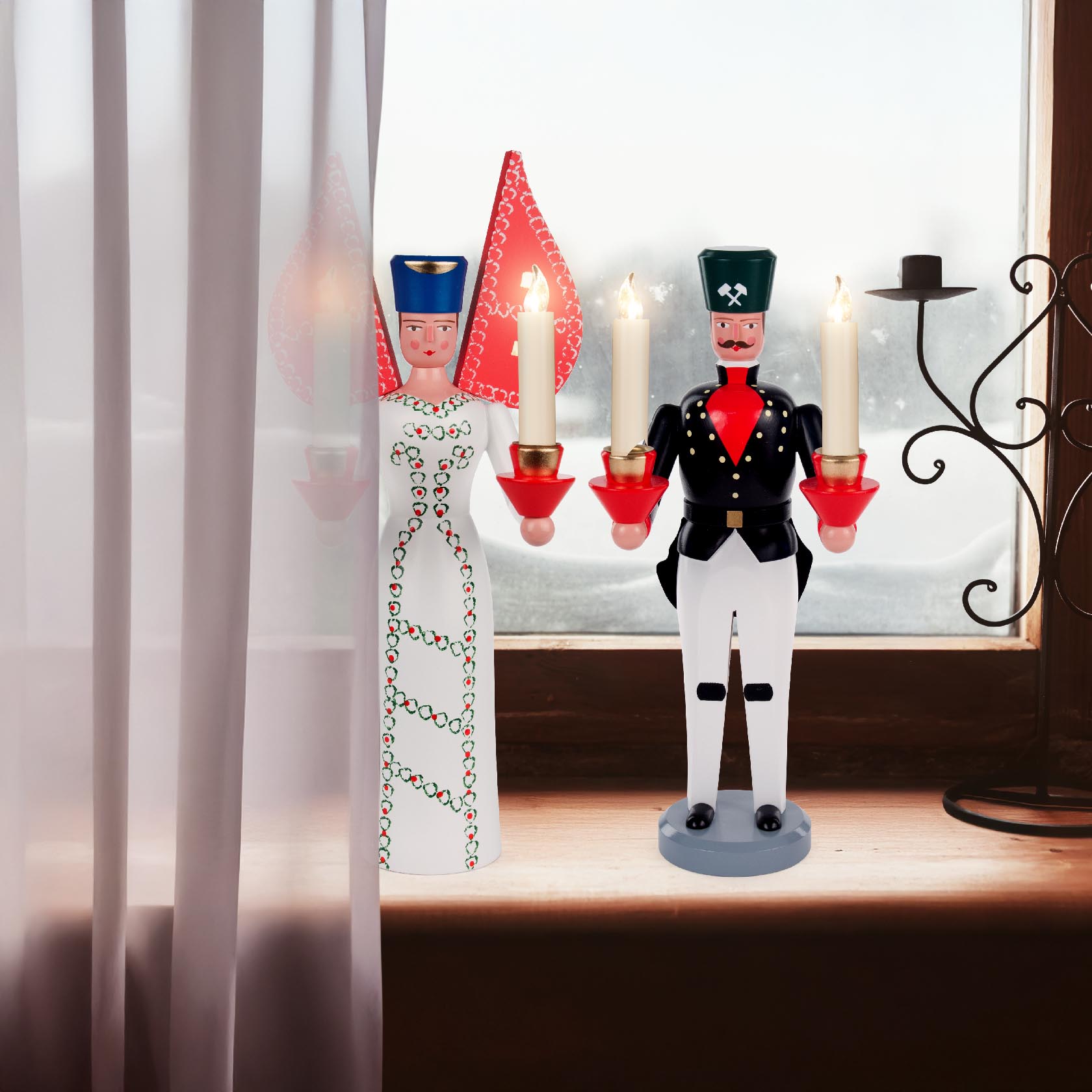 Two wooden candle holders in the shape of traditional figures, an angel and a soldier, each holding two lit candles, displayed on a windowsill.