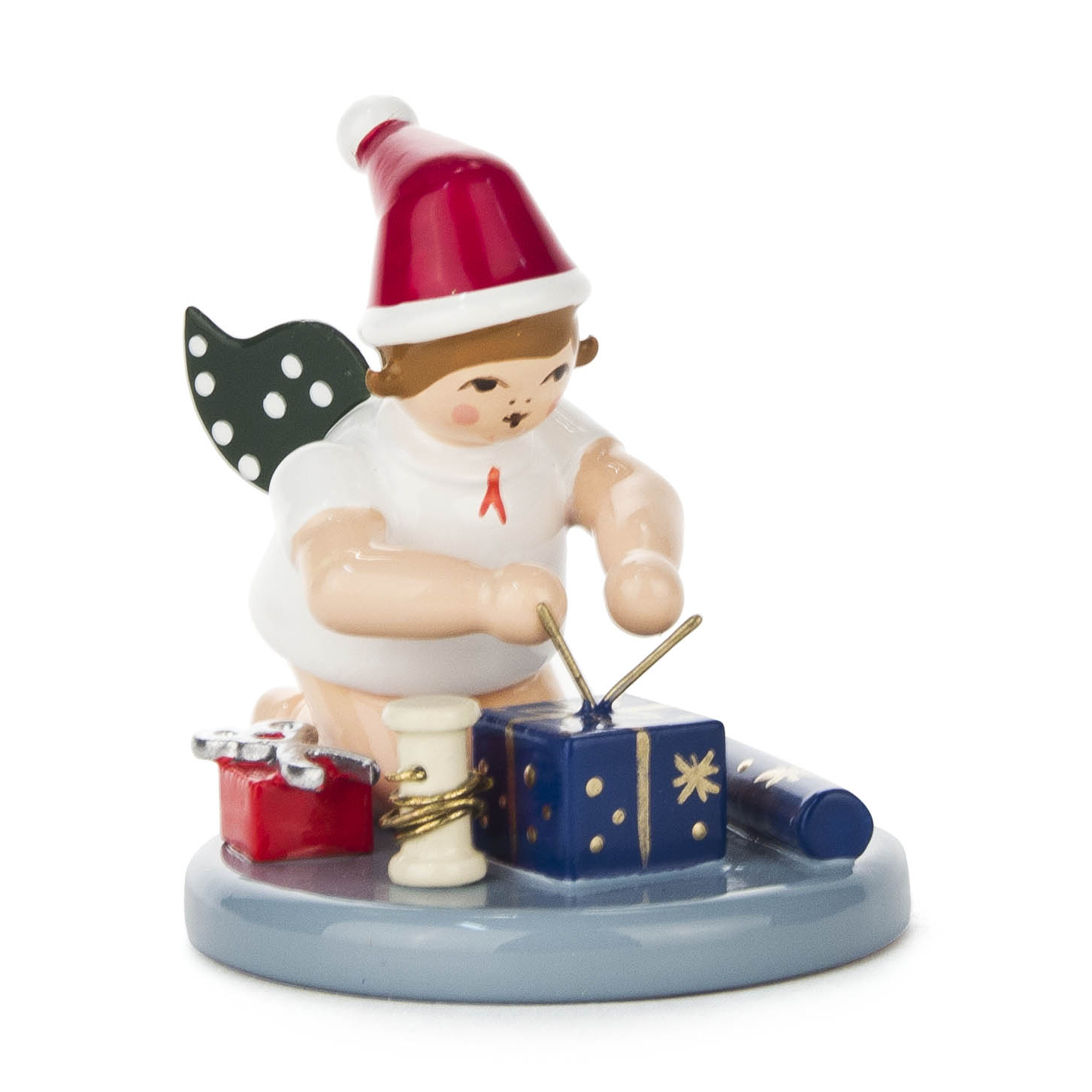 A kneeling angel figurine with wings and a Santa hat, wrapping a blue gift using ribbon, scissors, and wrapping paper, with a red gift nearby. im Dregeno Online Shop günstig kaufen