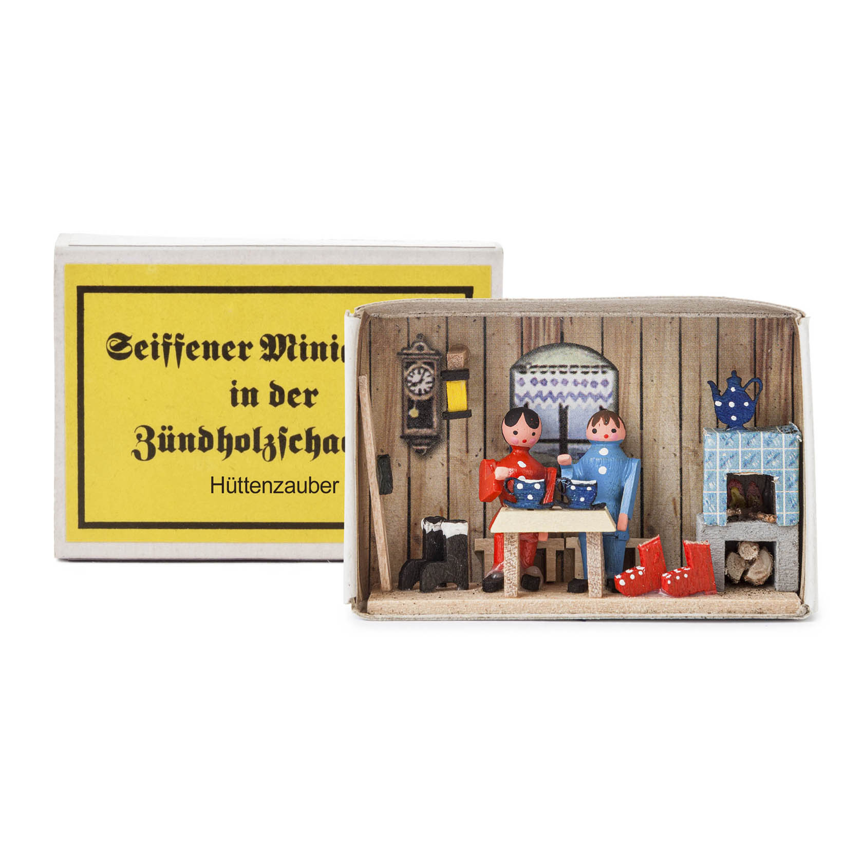 A matchbox diorama titled 'Hüttenzauber' with a small wooden interior scene featuring two painted wooden figures in traditional clothing with a chimney and a cuckoo clock. im Dregeno Online Shop günstig kaufen