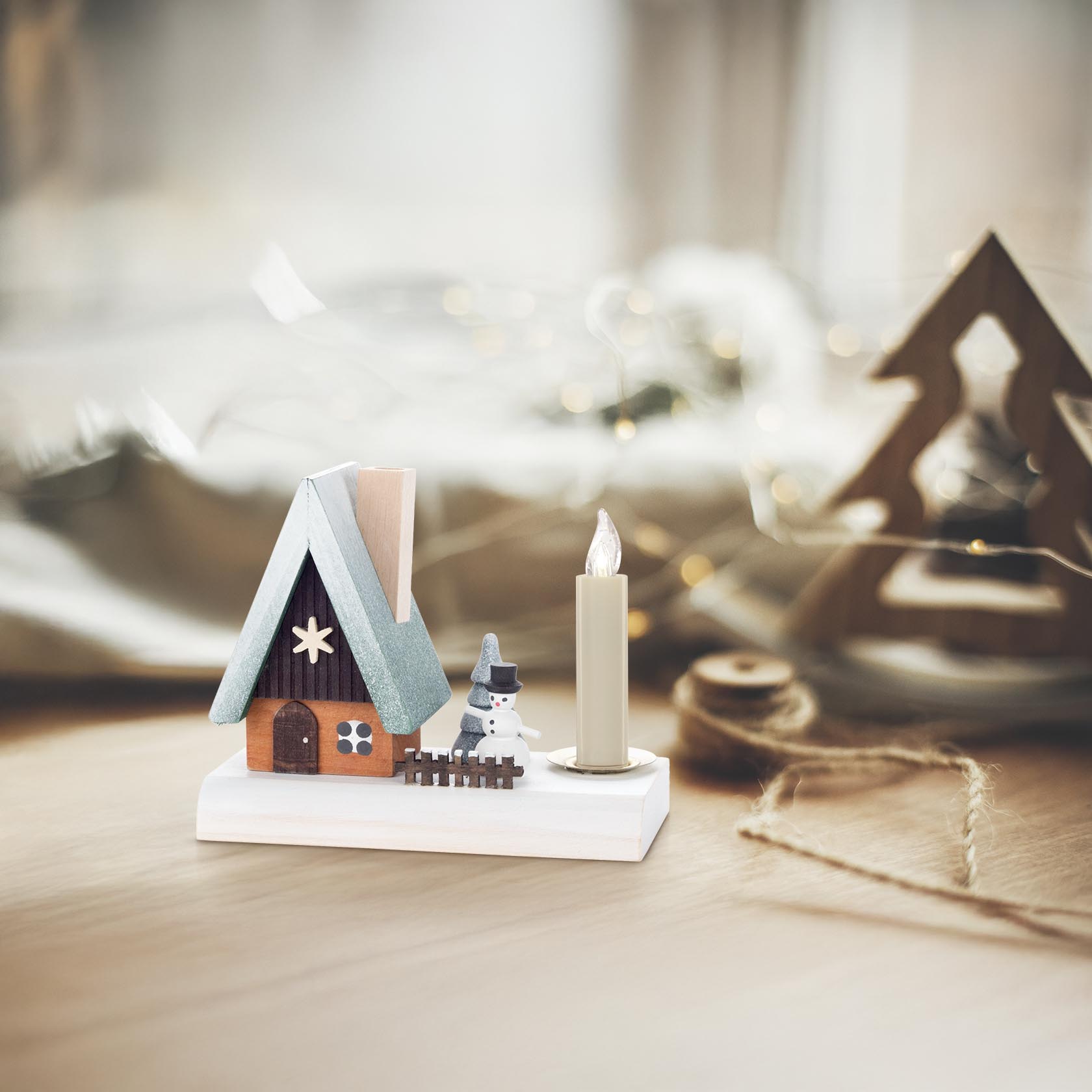 Kleines Holzräucherhaus mit blauem Dach und Schneemannfigur. Daneben eine angezündete Kerze. Im Hintergrund verschwommene Lichter.