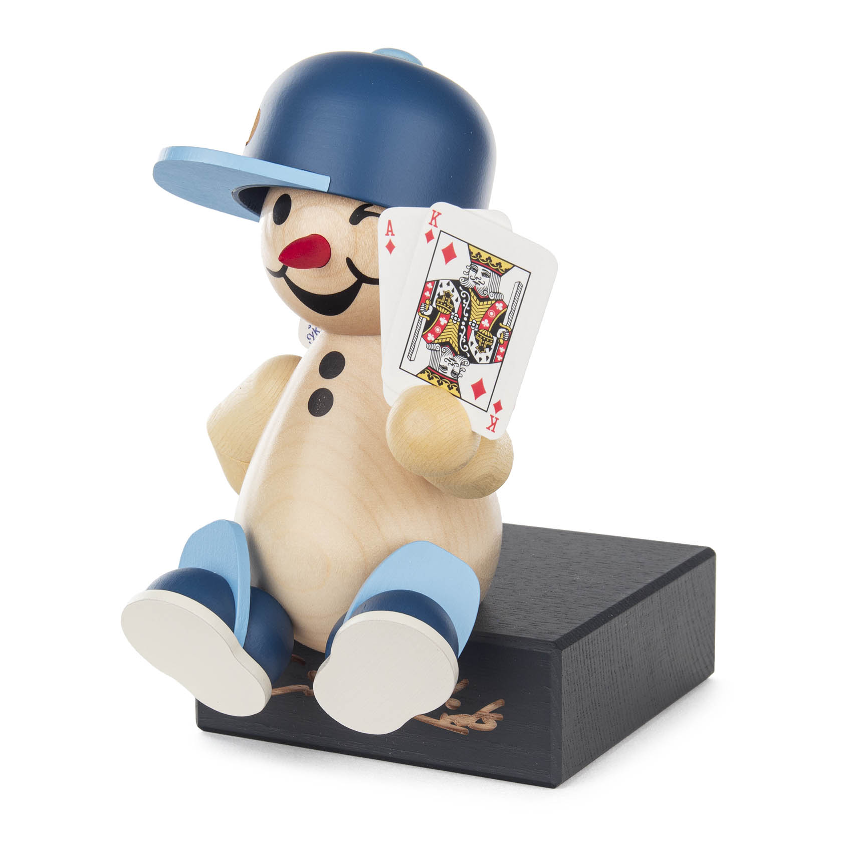 A wooden toy figure with a round body, sitting on a black platform. It wears a blue cap, red nose, and holds playing cards with a king of hearts visible.