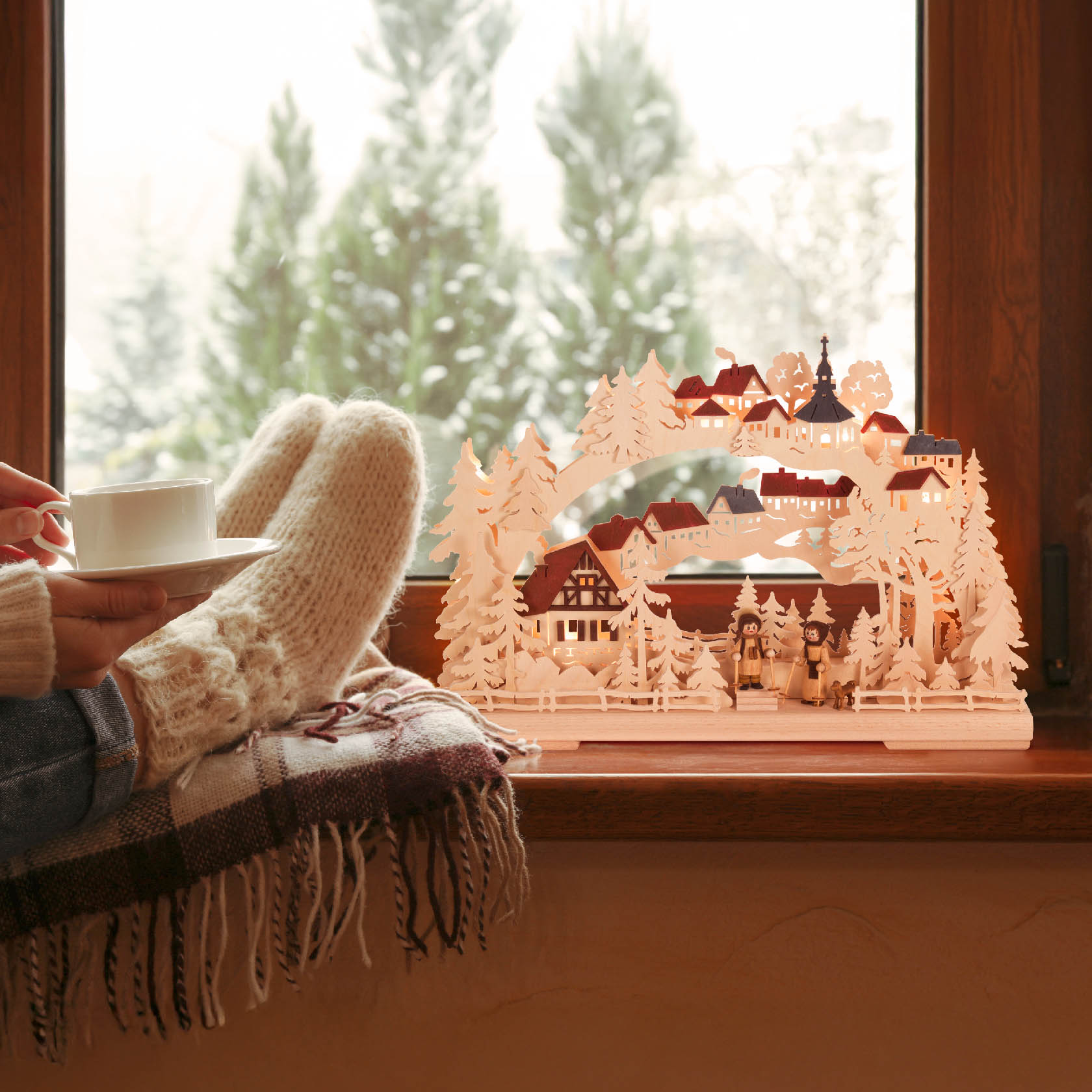 Ein Schwibbogen mit dem Motiv einer Winterwanderung, elektrisch beleuchtet, neben einer Person mit Wollsocken, die eine Tasse hält.