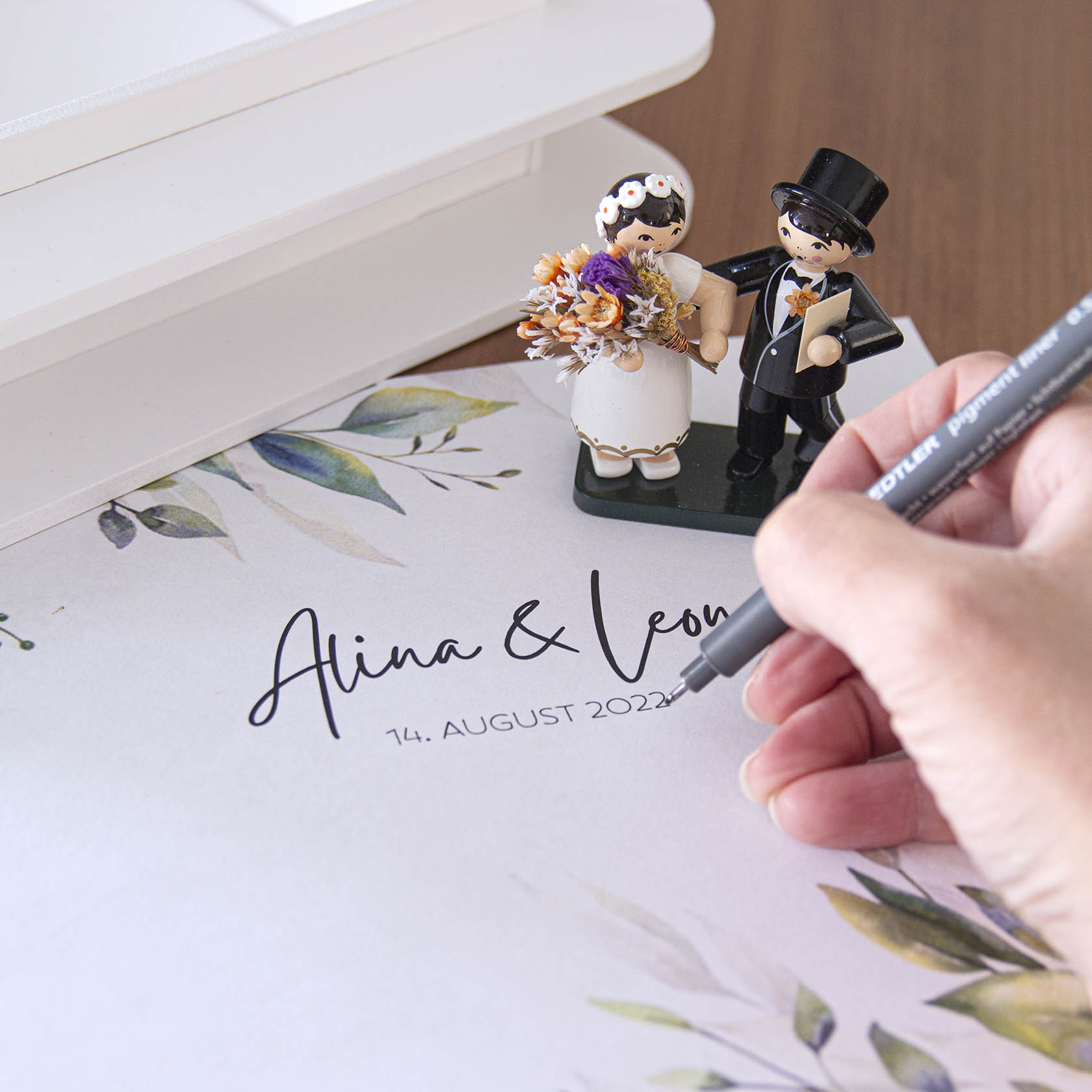 A close-up of a hand writing on a wedding card with bride and groom figurines holding flowers in the background.
