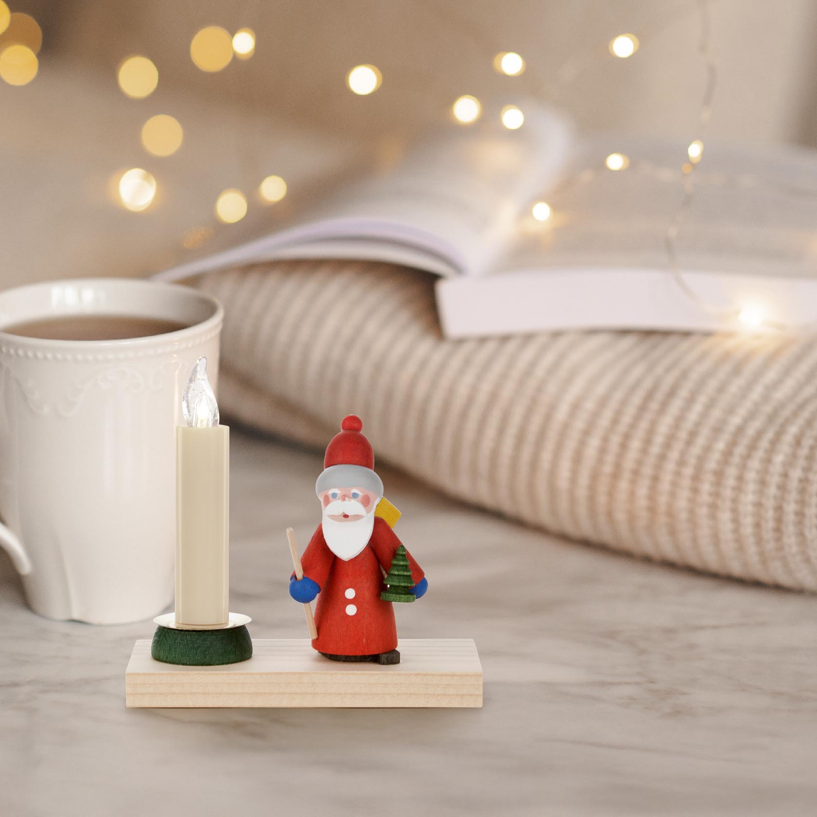 Festive candle holder with a wooden Santa figurine dressed in red, holding a green Christmas tree, next to a large white candle on a wooden base.