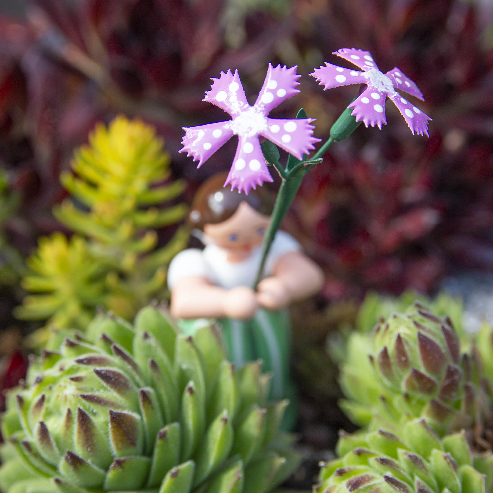 Eine Spielfigur eines Blumenmädchens hält eine violette Wiesennelke mit weißen Punkten in der Hand, umgeben von grünen Pflanzen.