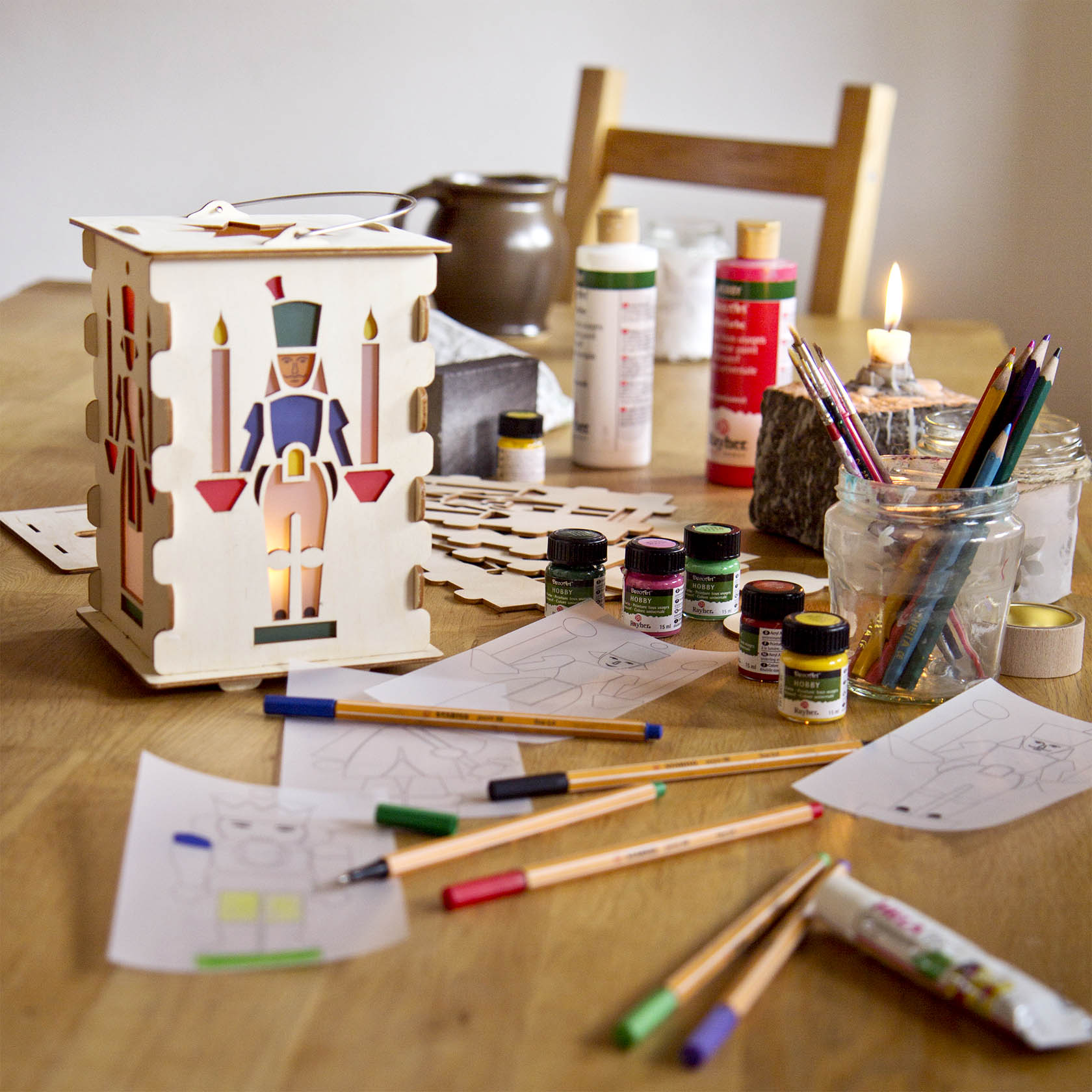 Craft set for a table lantern featuring a wooden nutcracker in the center, surrounded by paint bottles, colored pencils, and markers on a table.