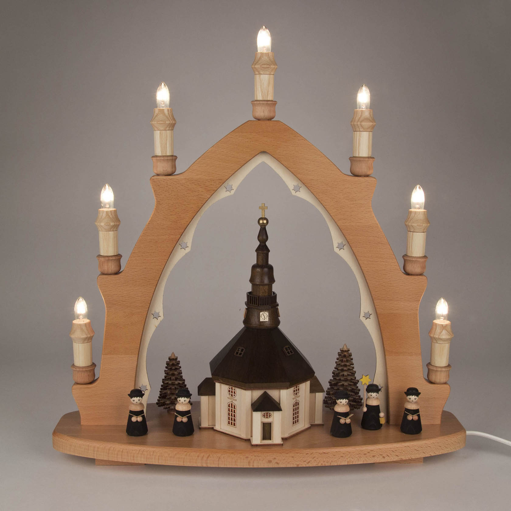 Schwibbogen aus Holz mit beleuchteter Seiffener Kirche und vier Kurrendesängern. Umgeben von Bäumen, mit sieben elektrischen Kerzen auf dem Bogen.