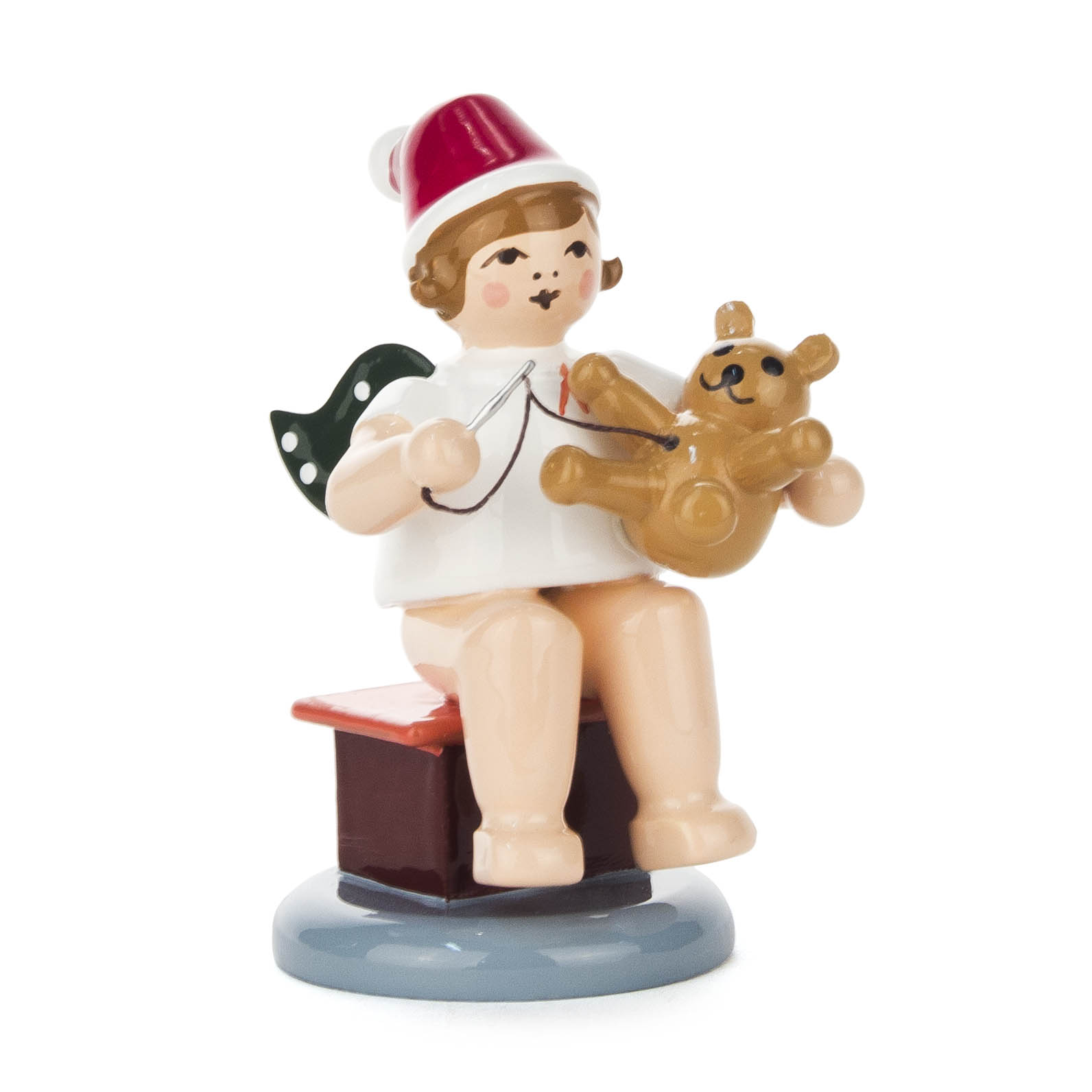 A wooden angel figurine sitting, holding a teddy bear and a sewing needle, wearing a red Santa hat and white outfit with green wings. im Dregeno Online Shop günstig kaufen