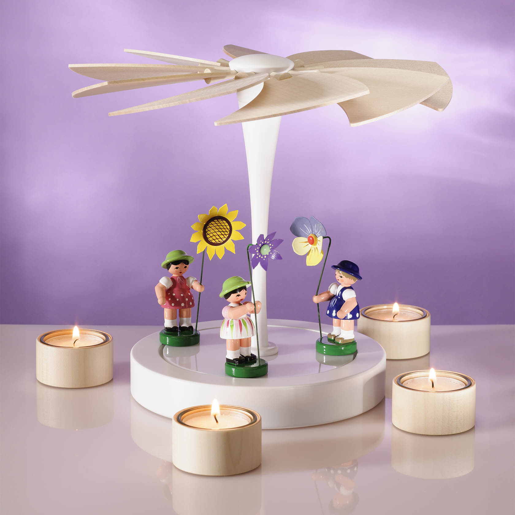 A white and natural wooden pyramid candle holder with four tea lights surrounding it. Features three tiny colorful figures carrying flowers.