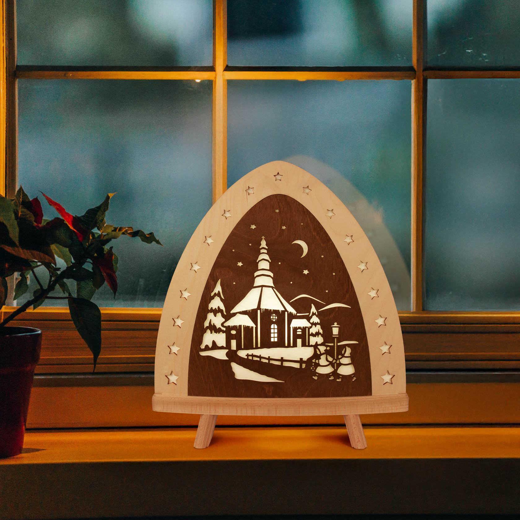 Wooden LED floor lamp featuring the silhouette of a church and snowy landscape with star cutouts, positioned on a windowsill.