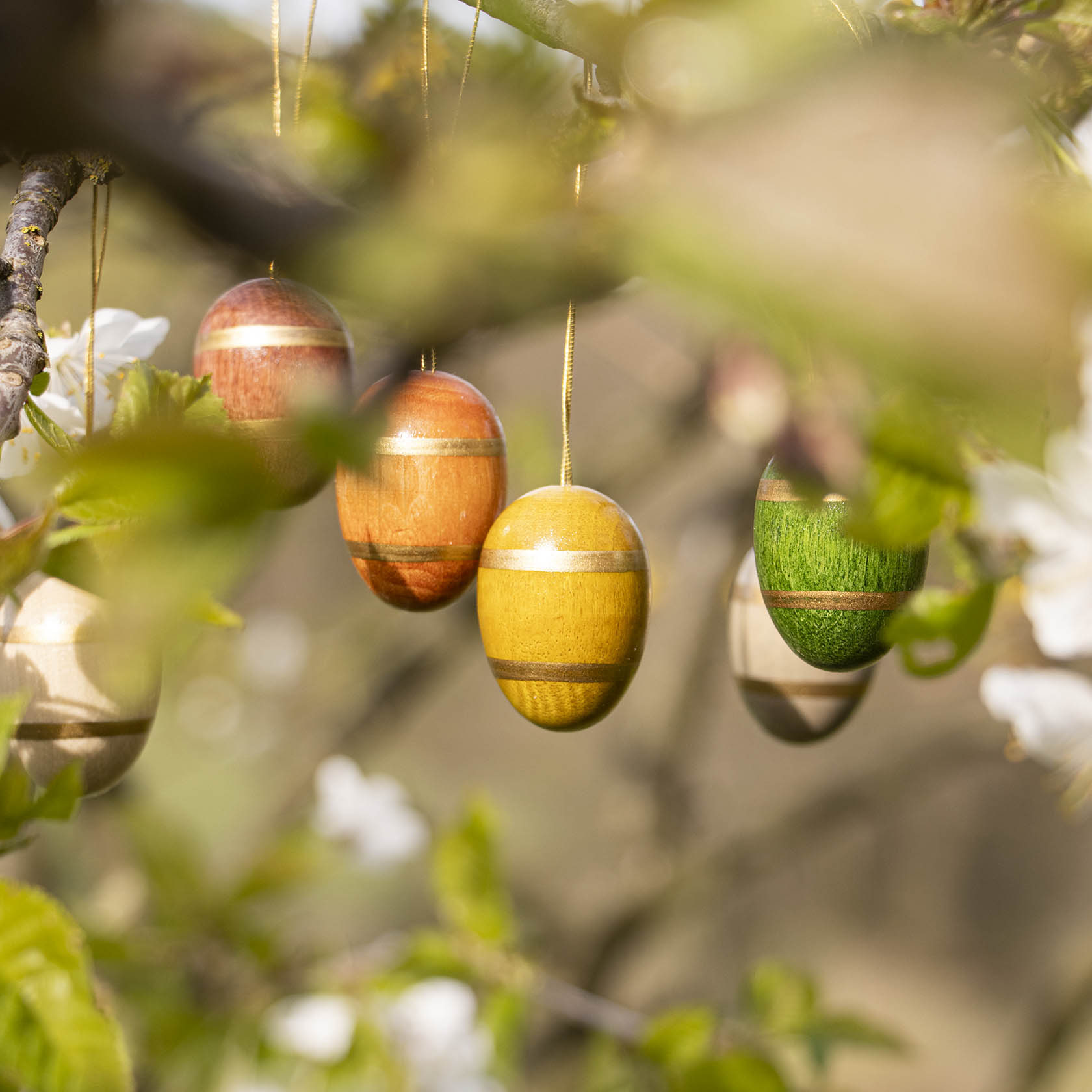 Fünf dekorative Ostereier in verschiedenen Farben mit horizontalen Streifen hängen an Zweigen in einem Garten.