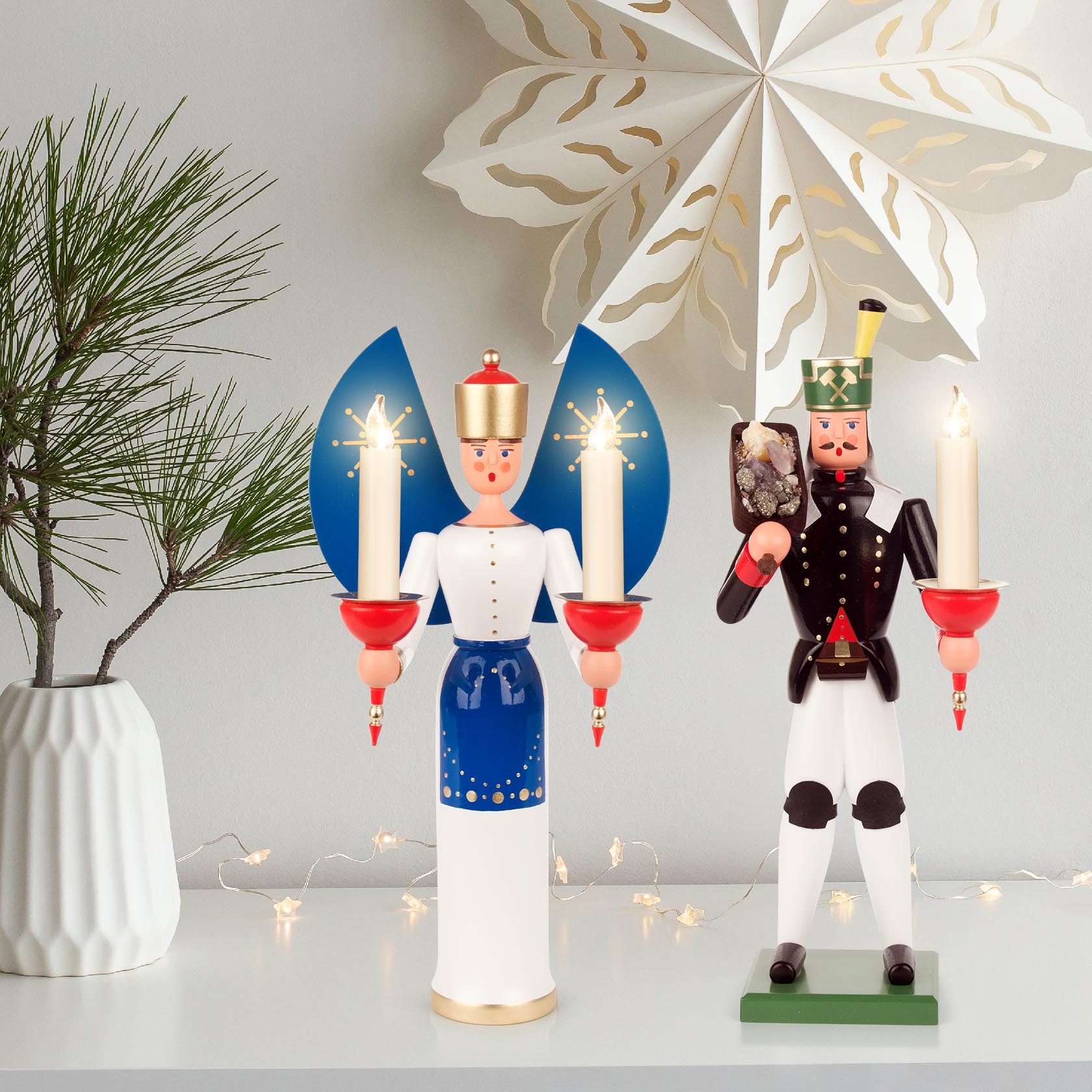 Zwei traditionelle Holzfiguren auf einem Tisch: links eine Engel-Figur mit blauen Flügeln, rechts ein Bergmann mit Erzstück und Kerzenhalter.