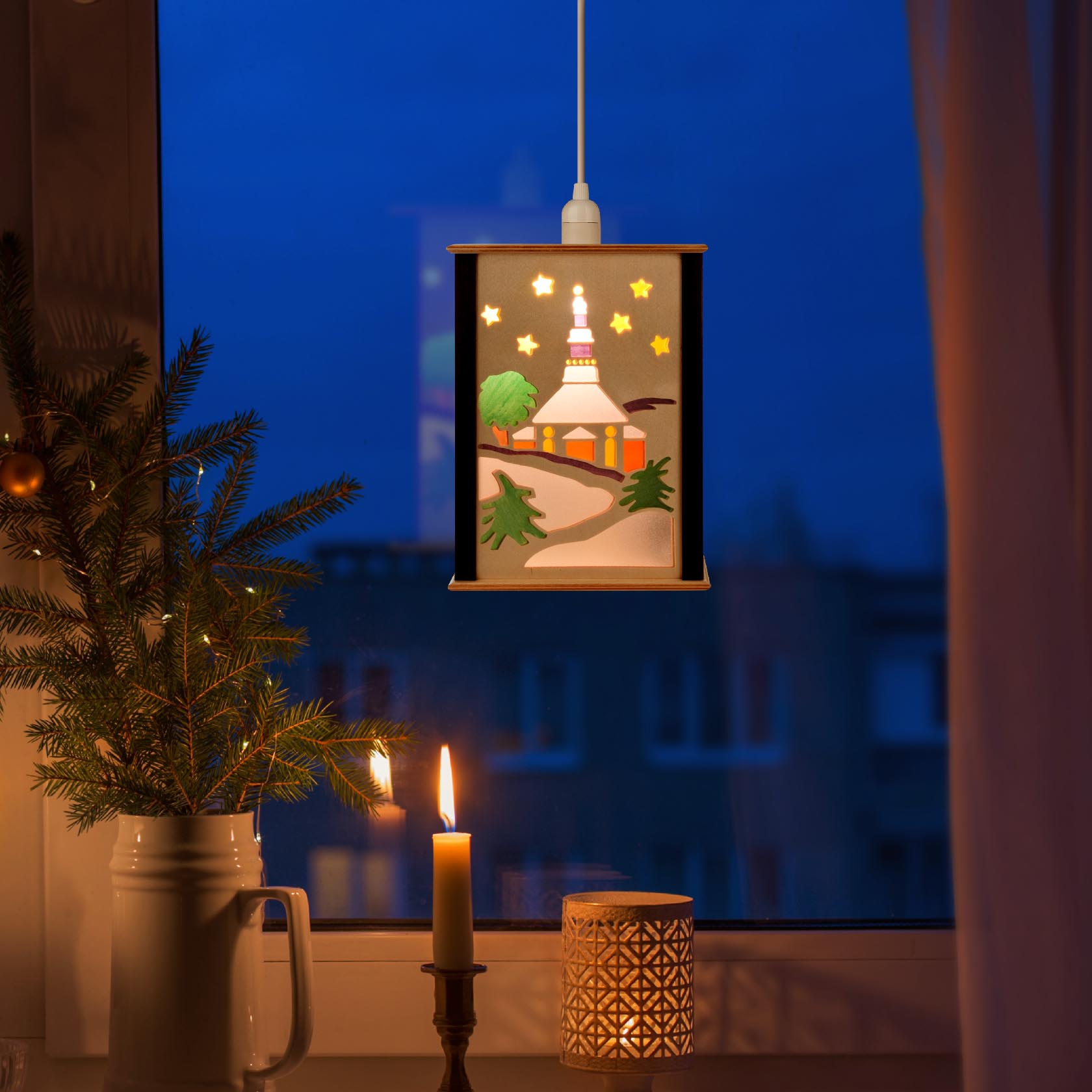 A small window lantern depicting the Seiffener Church and light angel, hung against a window. A candle and pine decorations are placed nearby.