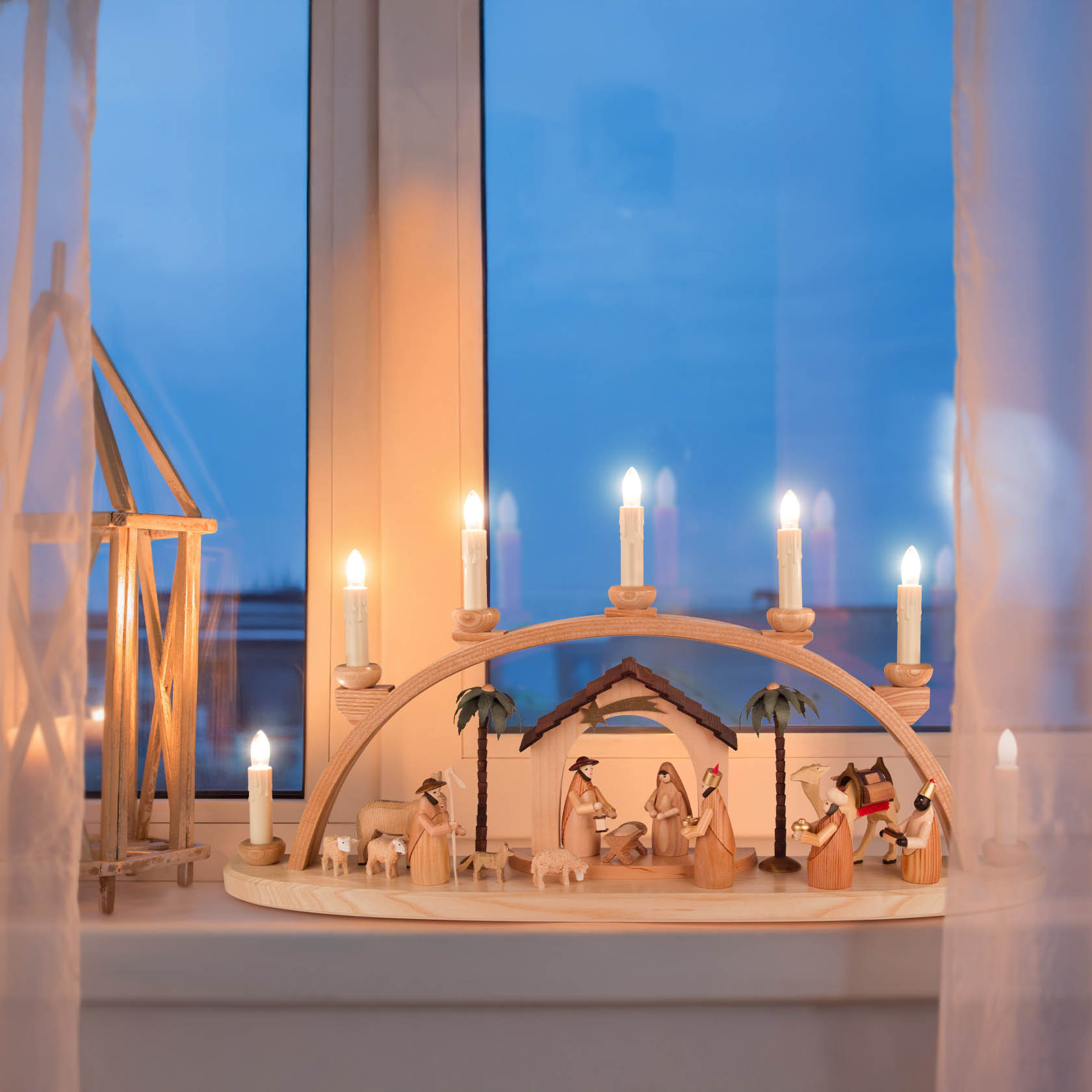 A natural wood Christmas arch (Schwibbogen) depicting the Nativity scene with electric candles illuminated in a window setting.