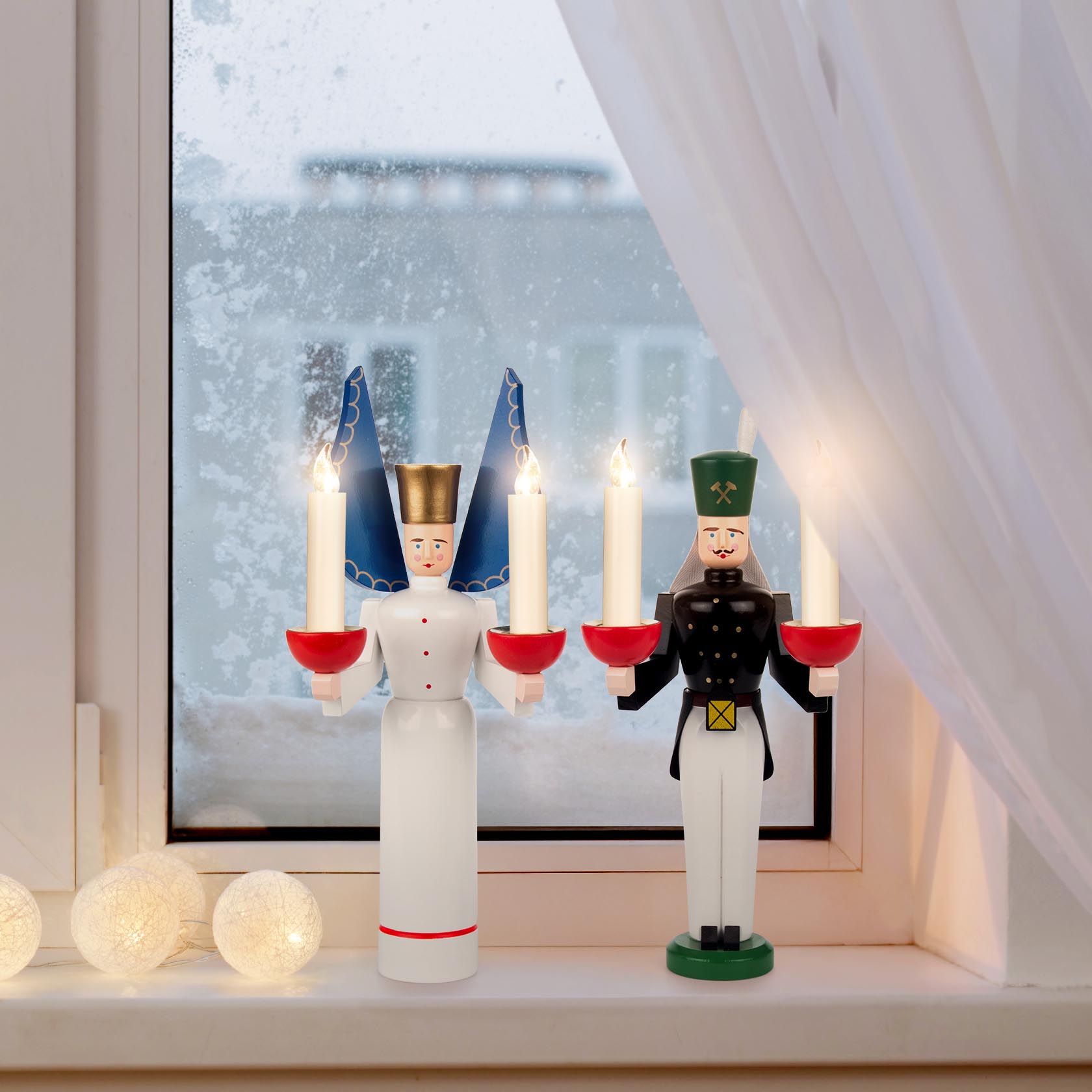 Ein traditionell gestalteter Engel und Bergmann aus Holz, halten jeweils Kerzenhalter mit brennenden Kerzen in den Händen, auf einer Fensterbank.